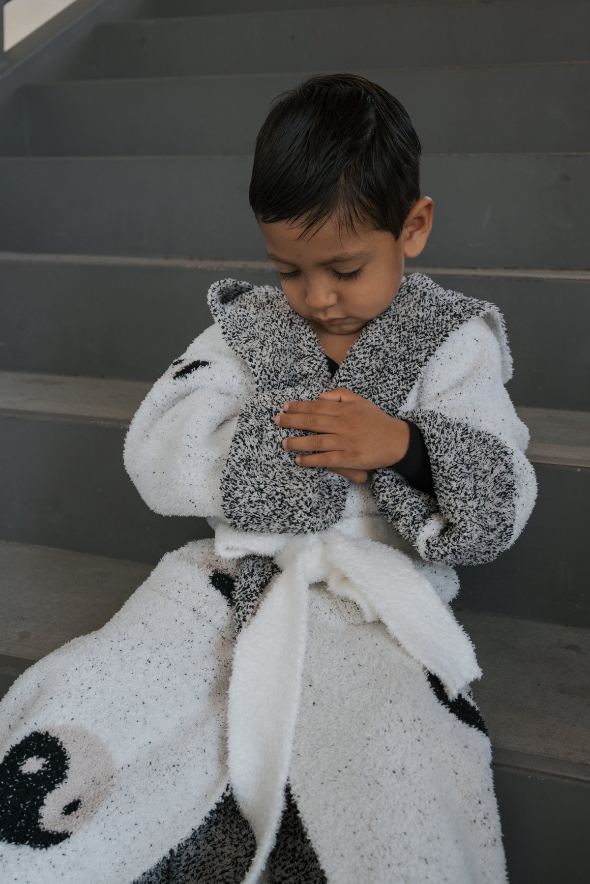 A young child sits on concrete stairs wearing a plush white forever french baby Children’s Robe | Yin Yang with a black yin yang pattern.
