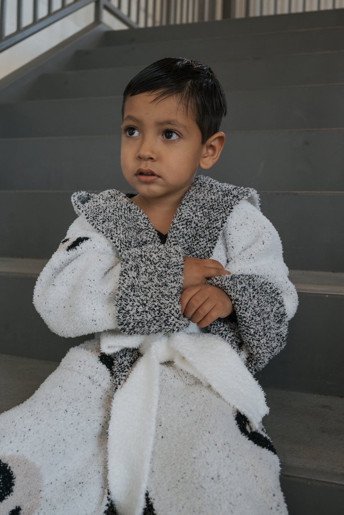 A young child sits on gray stairs wearing a Children’s Robe | Yin Yang, a hooded robe made of textured microfiber feather yarn.