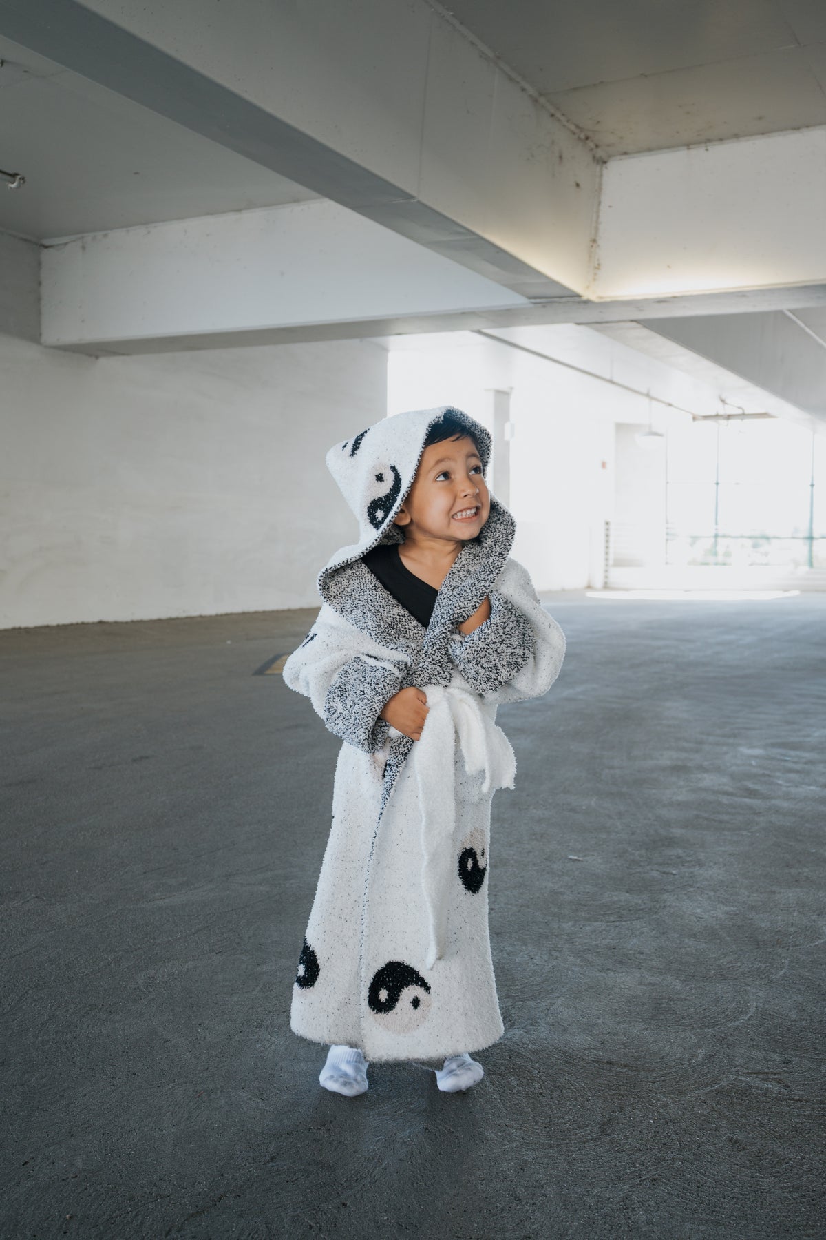 A young child wearing a white hooded Children’s Robe | Yin Yang with a black yin-yang pattern, standing in a parking garage.