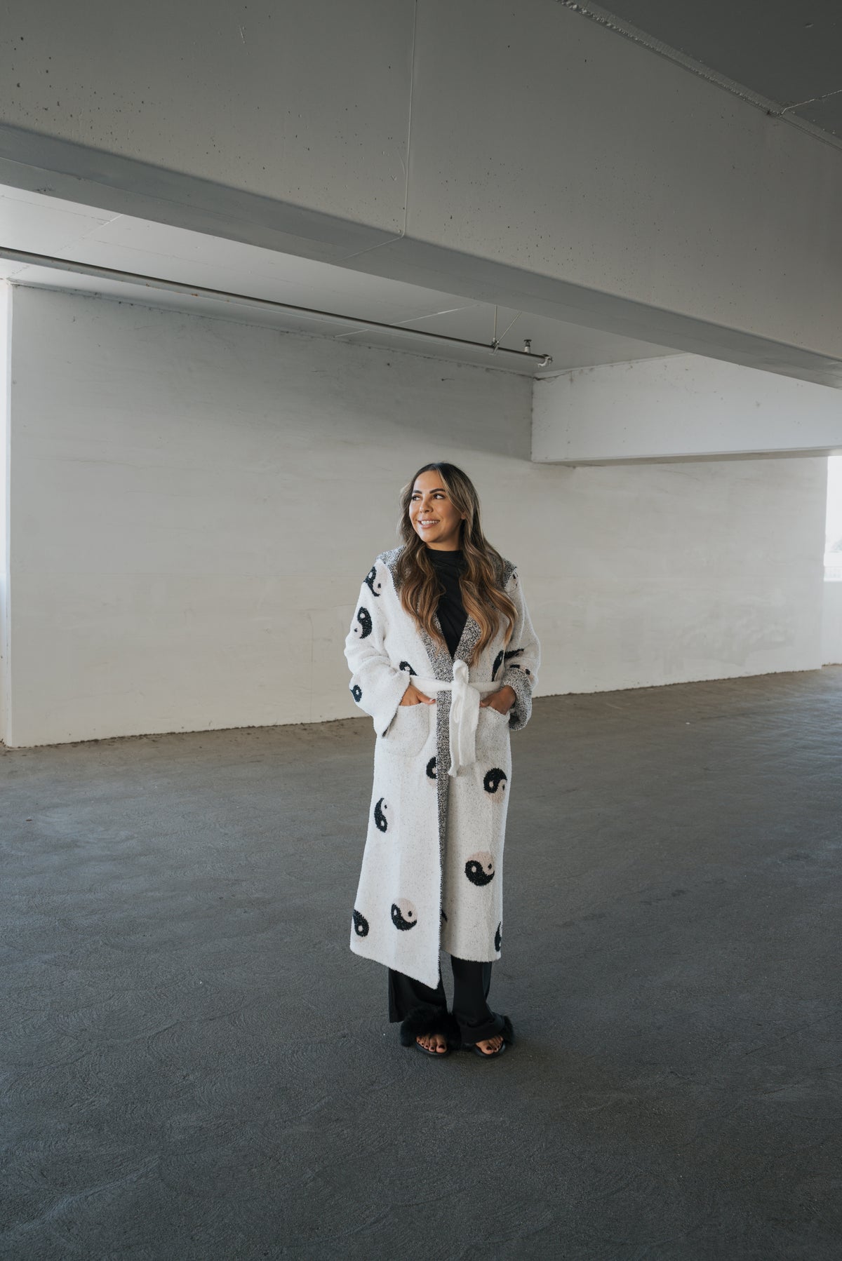 An adult wears a forever french baby Adult Robe | Yin Yang, a long, white, microfiber robe patterned with black and brown yin-yang symbols, while standing in a parking garage.