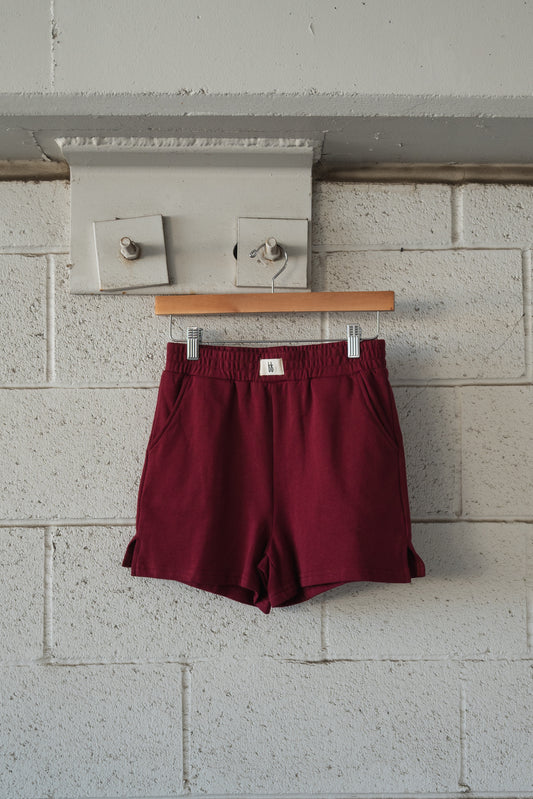 Forever French Baby Women’s Terry Shorts in deep cranberry with an elastic waistband, hanging on a wooden hanger against a white brick wall.