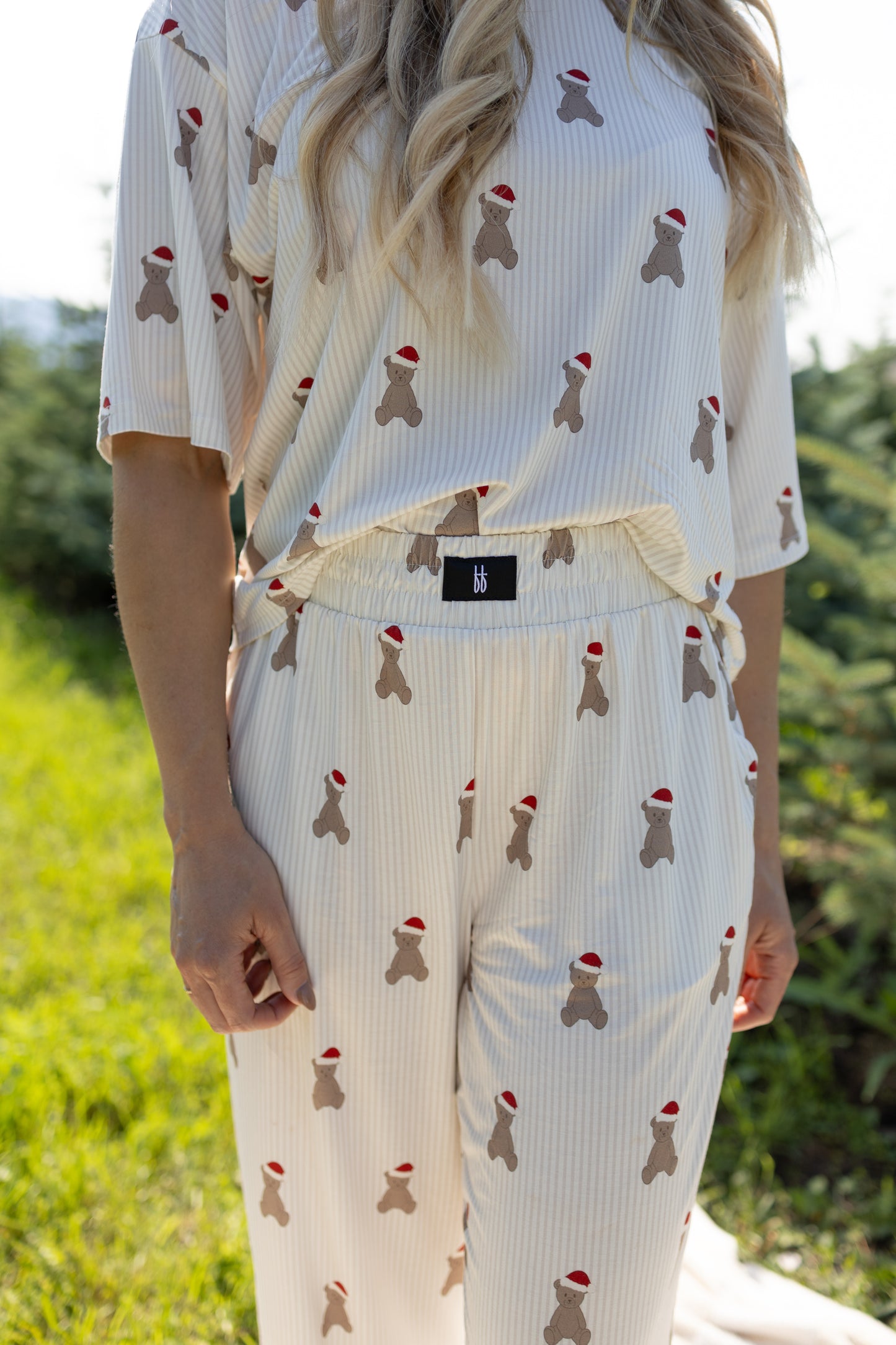 A woman stands outdoors on green grass, wearing the forever french baby Women's Boxy Tee Lounge Set | Santa Teddy—a cream-colored, breathable bamboo pajama set featuring small teddy bear prints in red Santa hats. Only her torso and legs are visible.
