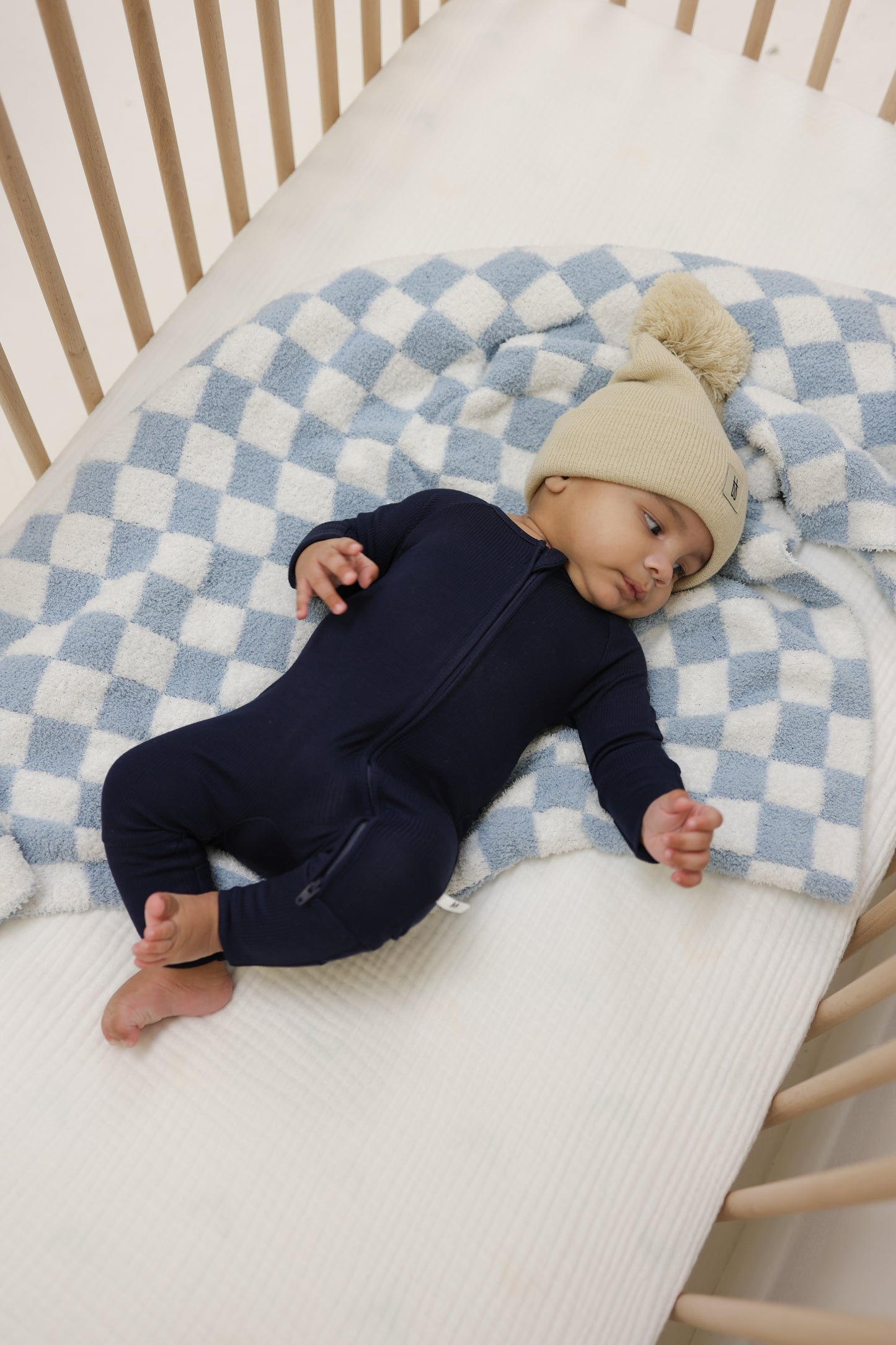 A baby in forever french baby's Bamboo Zip Pajamas | Blueberry Ribbed and a beige knit hat with pom-pom lies on a blue and white checkered blanket in a wooden crib.