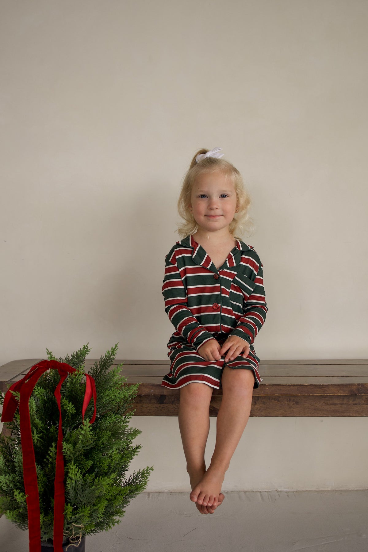 A young girl wears a forever french baby Bamboo Sleeping Dress | Holiday Stripe while sitting on a wooden bench next to a small potted tree.