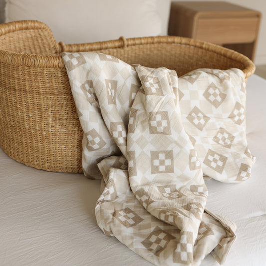 A woven wicker bassinet with the forever french baby Muslin Quilt | Cottage Quilt, featuring a beige and white geometric muslin cotton design, draped over its edge—perfect for stylish baby nursery bedding.
