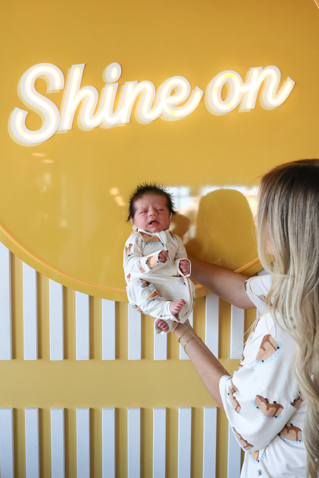 A blonde woman holds her newborn in front of a yellow wall with "Shine on," both wearing matching forever french baby Bamboo Zip Pajamas | Sunday Stack, enjoying the comfort and breathability of their sleepwear.