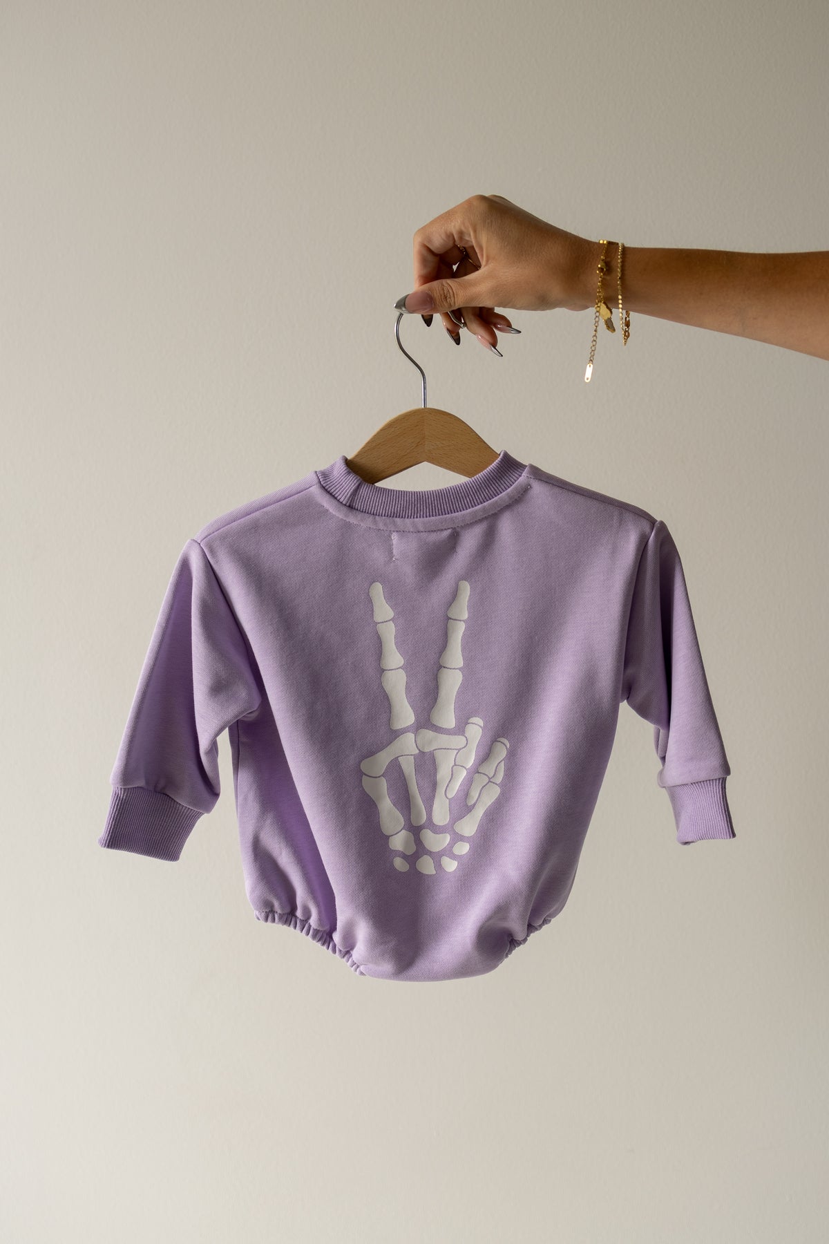 A hand holds a hanger displaying the Sweatshirt Romper | Enchanted Skelly by forever french baby, made from lavender 100% cotton and featuring a white skeleton hand making a peace sign on the back—an ideal pick for fans of the Midnight Skelly style.