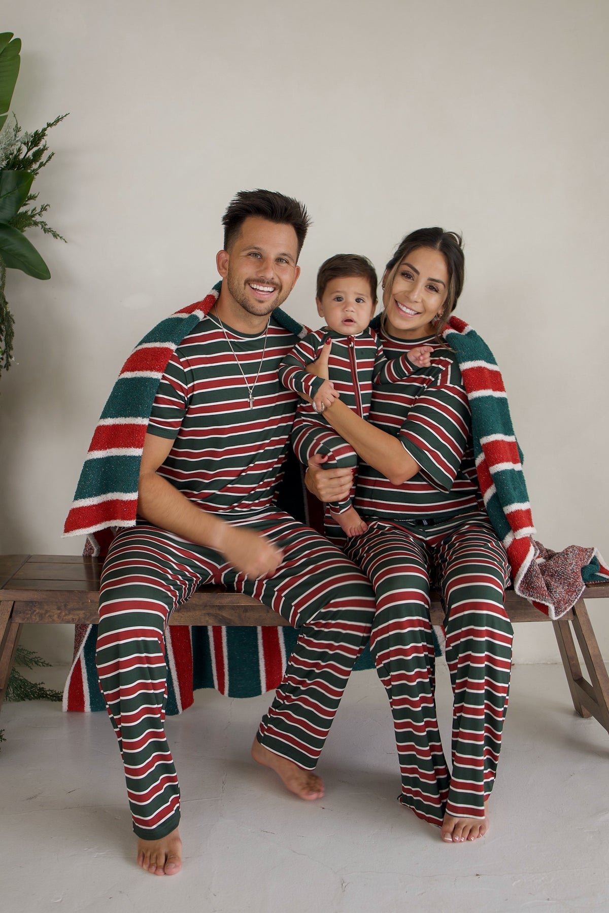 A family of three sits on a bench in matching holiday stripe pajamas, wrapped in a plush, Oeko-Tex certified *PRE-ORDER* Plush Blanket | Holiday Stripe with the same pattern.