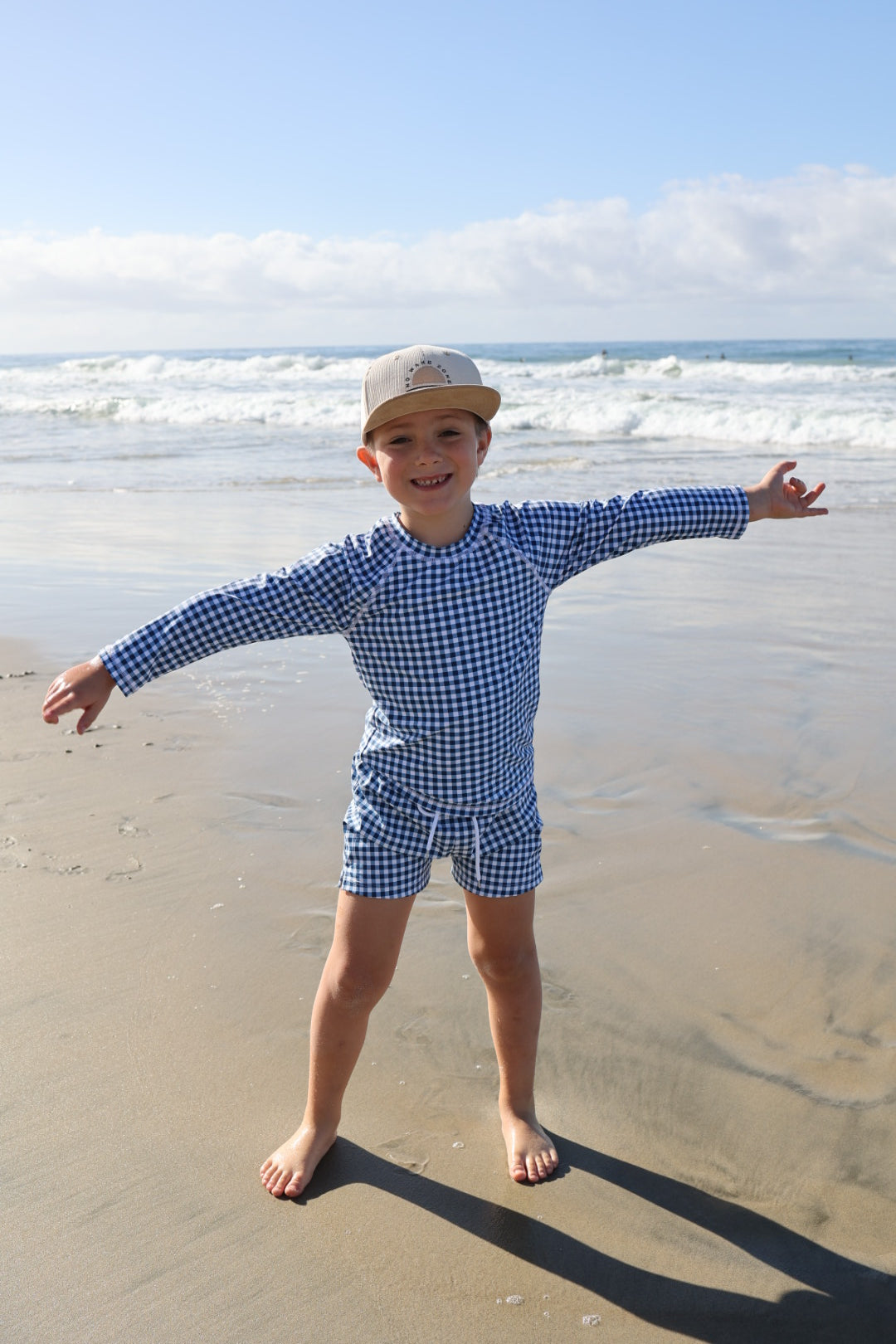 A young child wears forever french baby's Blue Gingham Child Board Short and a beige cap, smiling barefoot on the beach with arms outstretched. Waves and a cloudy sky create the perfect backdrop for this summer look.