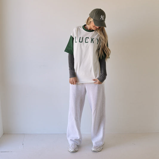 A person in a green cap, white "Adult Cotton Tee | Lucky" by forever french baby from the Lucky Collection, green sleeves under the tee, and white sweatpants stands indoors looking down, long hair partly covering their face.