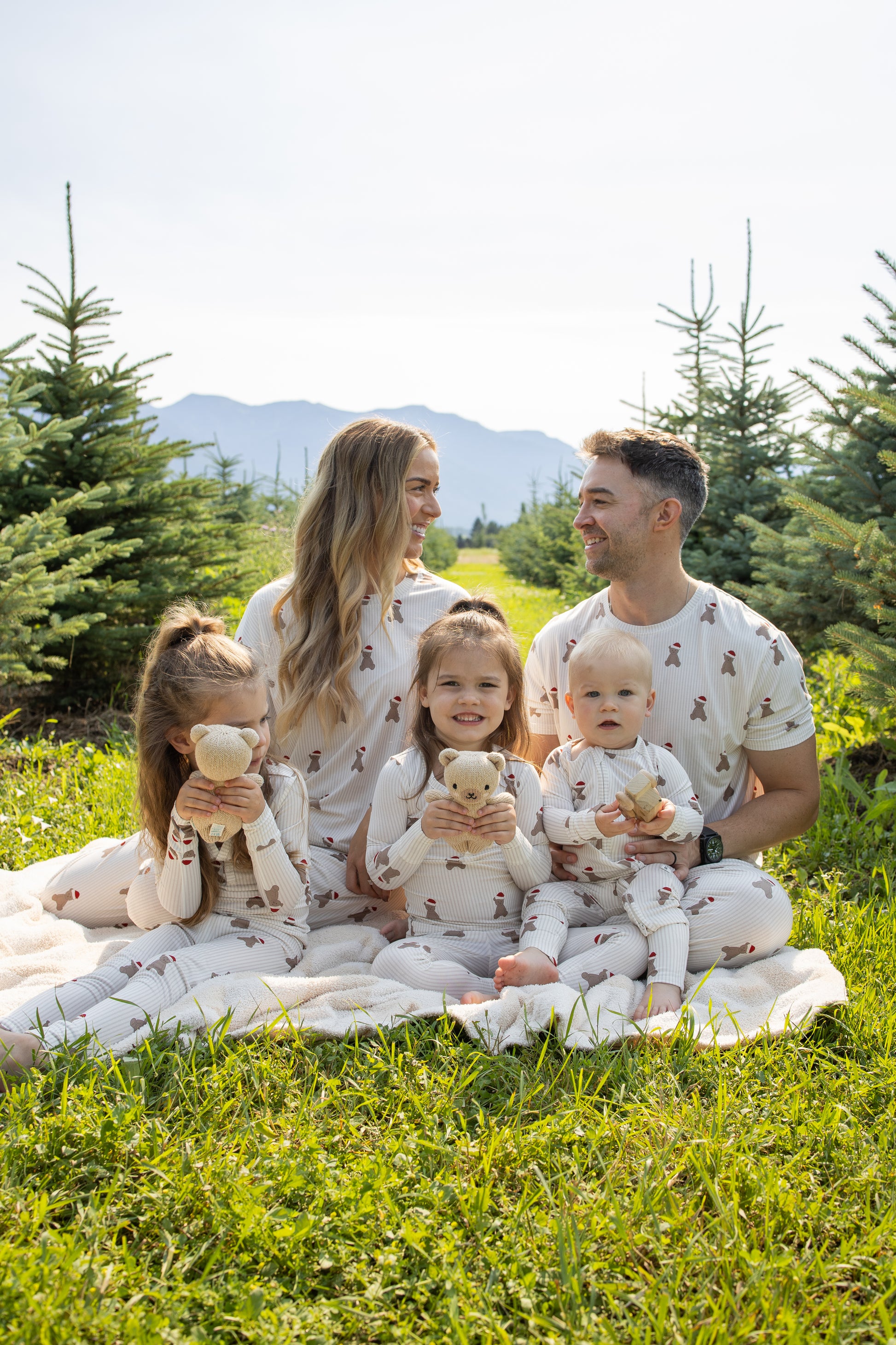 A family of four enjoys the outdoors among small evergreen trees, all dressed in forever french baby Bamboo Two Piece Pajamas | Santa Teddy. The parents share a smile while their children hold Santa Teddy stuffed animals and grin at the camera.