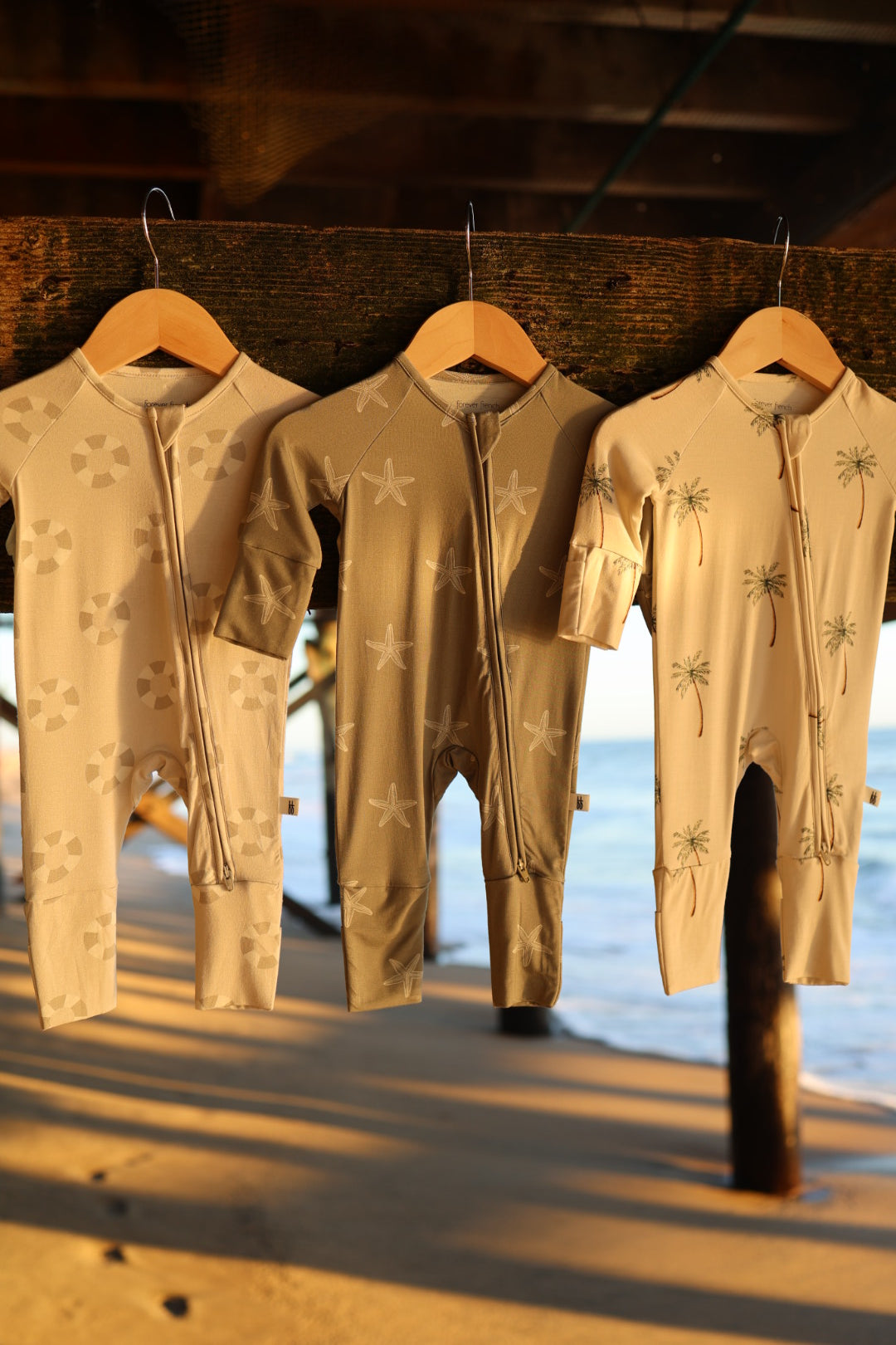Three beige Bamboo Zip Pajamas by forever french baby, made from soft, hypoallergenic fabric, display geometric shapes, starfish, and palm trees as they hang on wooden hangers under a sunlit pier with the beach behind.