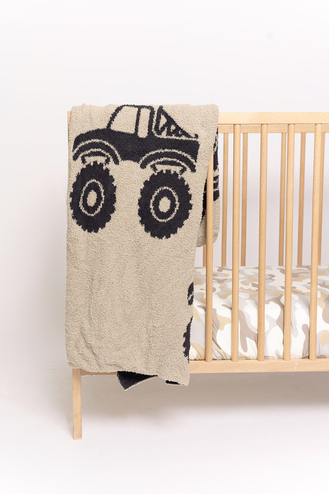 A forever french baby Plush Blanket | Monster Truck in beige, Oeko-Tex certified, is draped over a light wooden crib with a patterned mattress. The background is plain white.