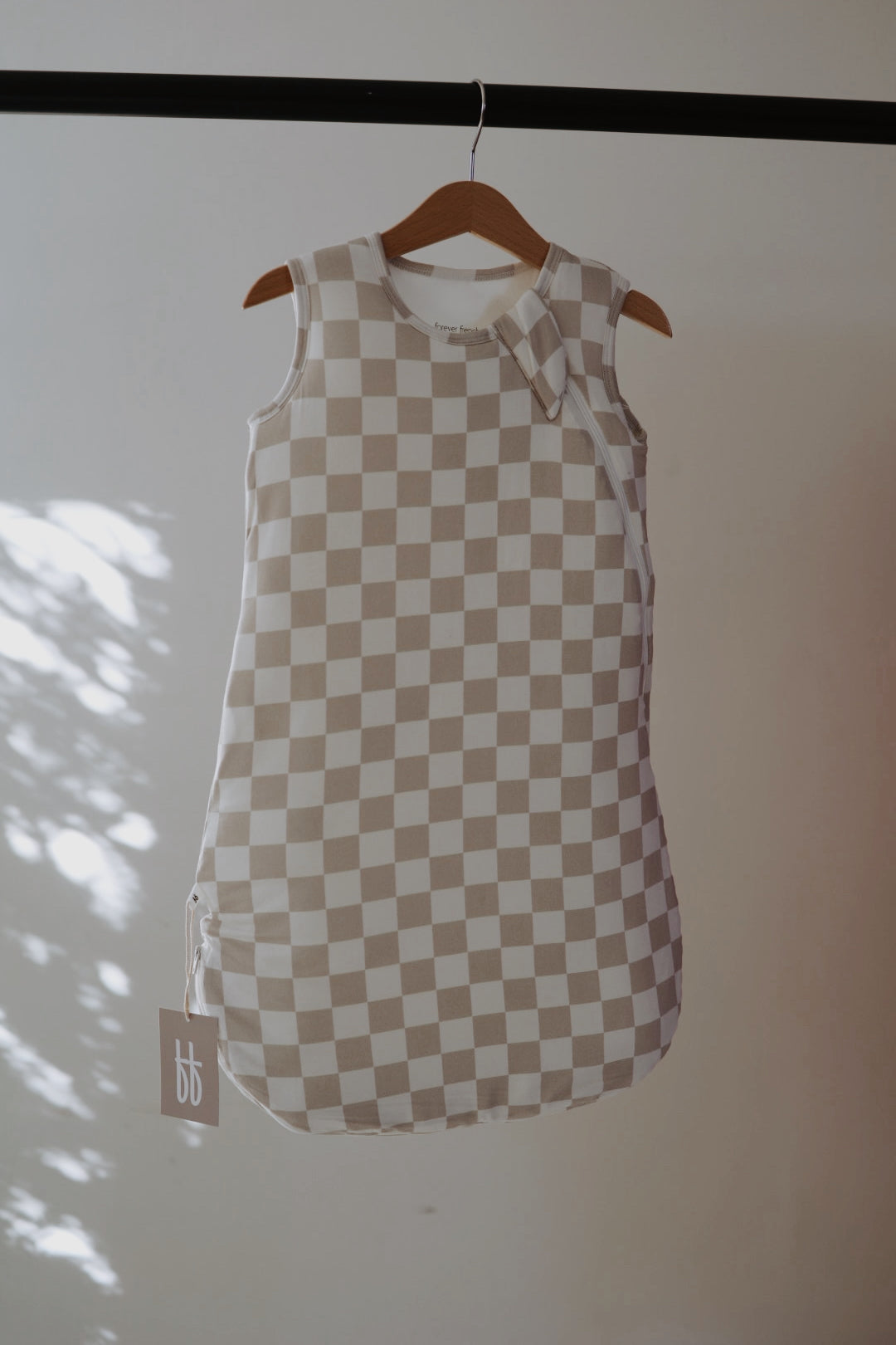 The Bamboo Sleep Sack | Checkerboard from forever french baby, featuring a beige and white checkerboard print, hangs on a wooden hanger as sunlight casts soft shadows. A "bb" tag accents this sleeveless, cozy wearable blanket.