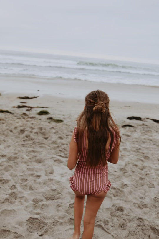 A young girl is walking towards the ocean on a sandy beach. She has long hair and is wearing a red and white striped "One Piece Girls Swim Suit | Boardwalk" by forever french baby, featuring earth tone stripes. The sky is overcast, and the waves are gently rolling in, offering a serene backdrop for her UPF 50+ protected adventure.