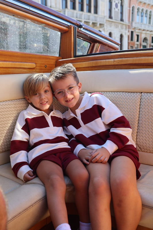 Two young boys wearing matching Child Rugby Sweatshirt | Deep Cranberry and shorts sit together on a boat seat.