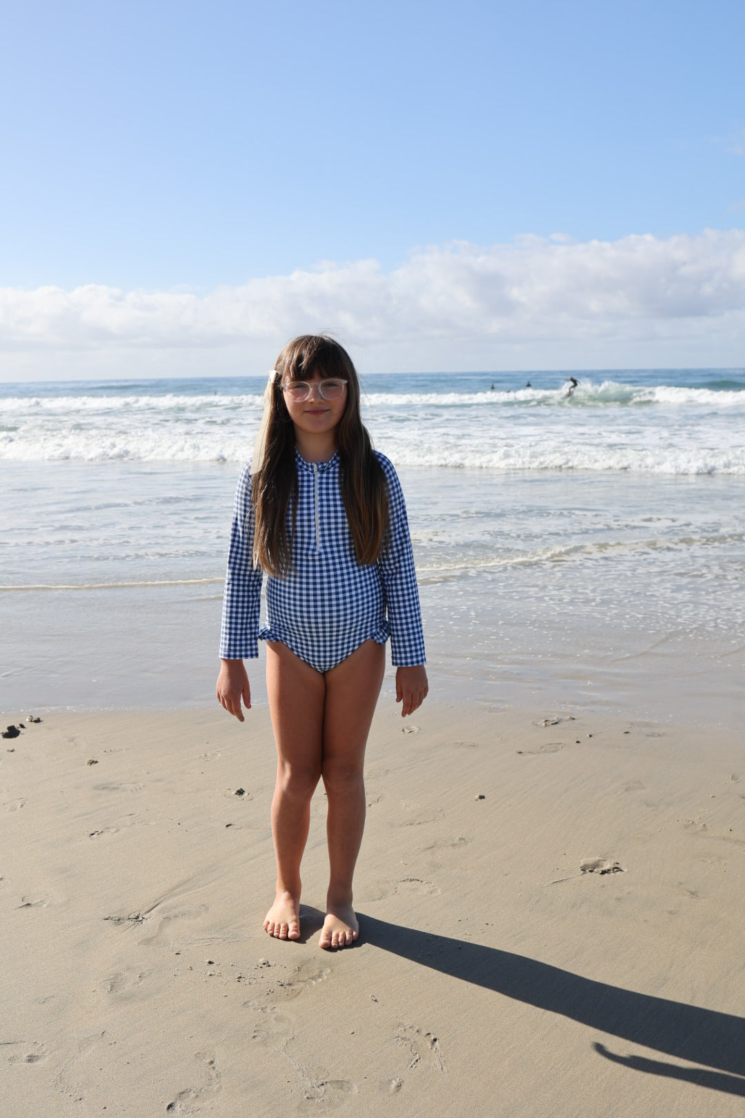 A girl stands on the beach in the forever french baby Girls Long Sleeve Swimsuit in Blue Gingham, ready for sunny adventures.