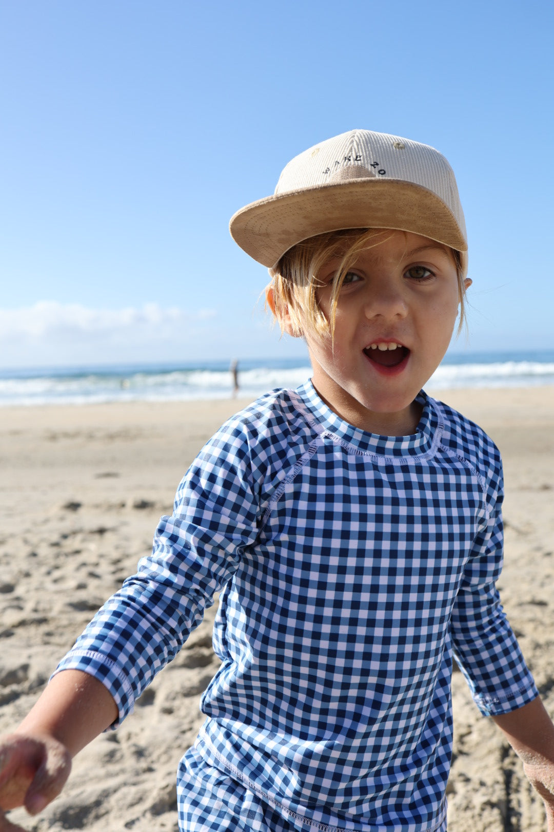 A young child in the forever french baby Child Rash Guard | Blue Gingham smiles on a sandy beach, with the ocean and blue sky in the background.