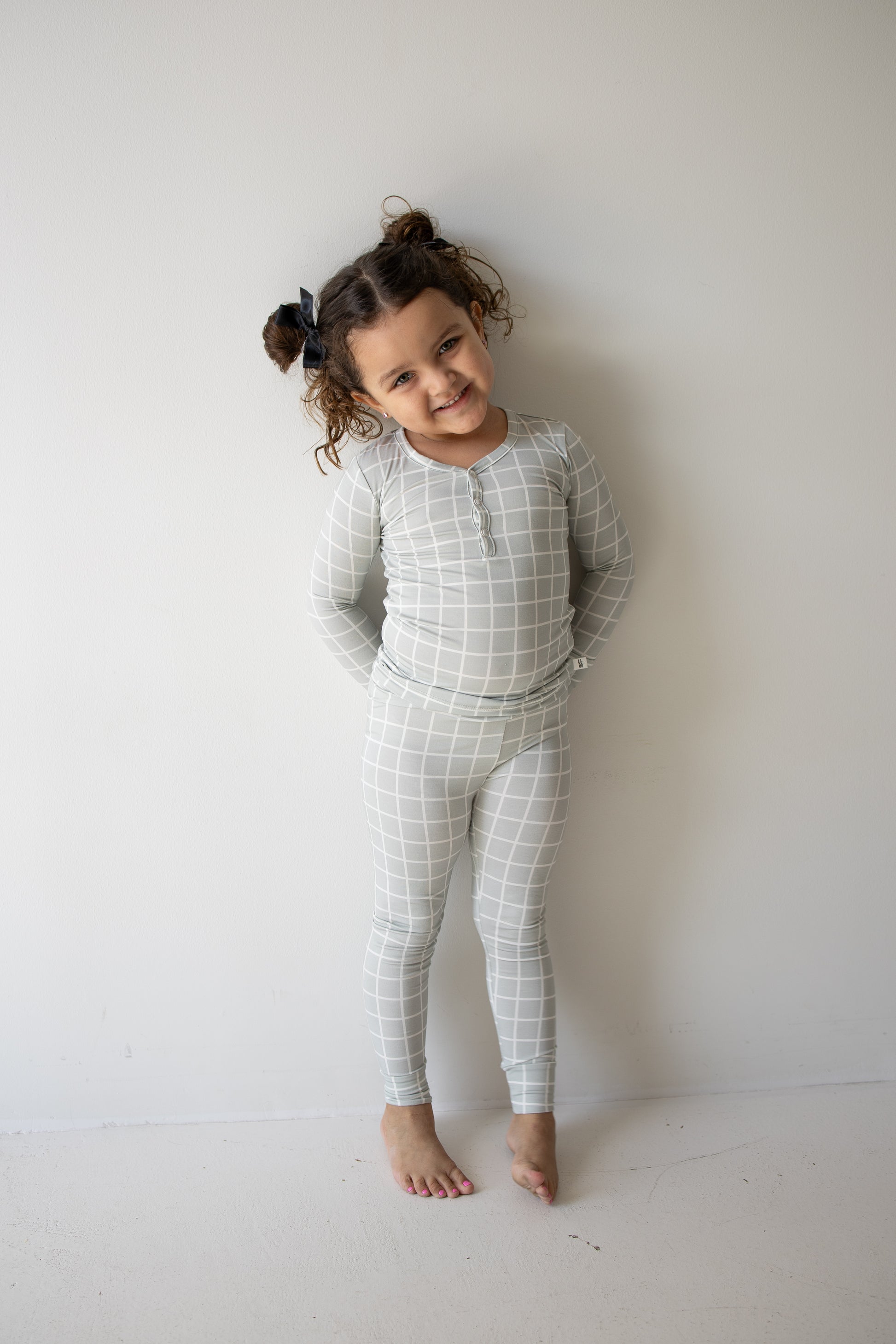 A smiling young child with curly hair and bows poses against a white wall, wearing the forever french baby Bamboo Two Piece Pajamas in Foggy Grid—a light gray long-sleeve set with matching leggings featuring a white grid pattern, perfect for all-day comfort in breathable bamboo fabric.