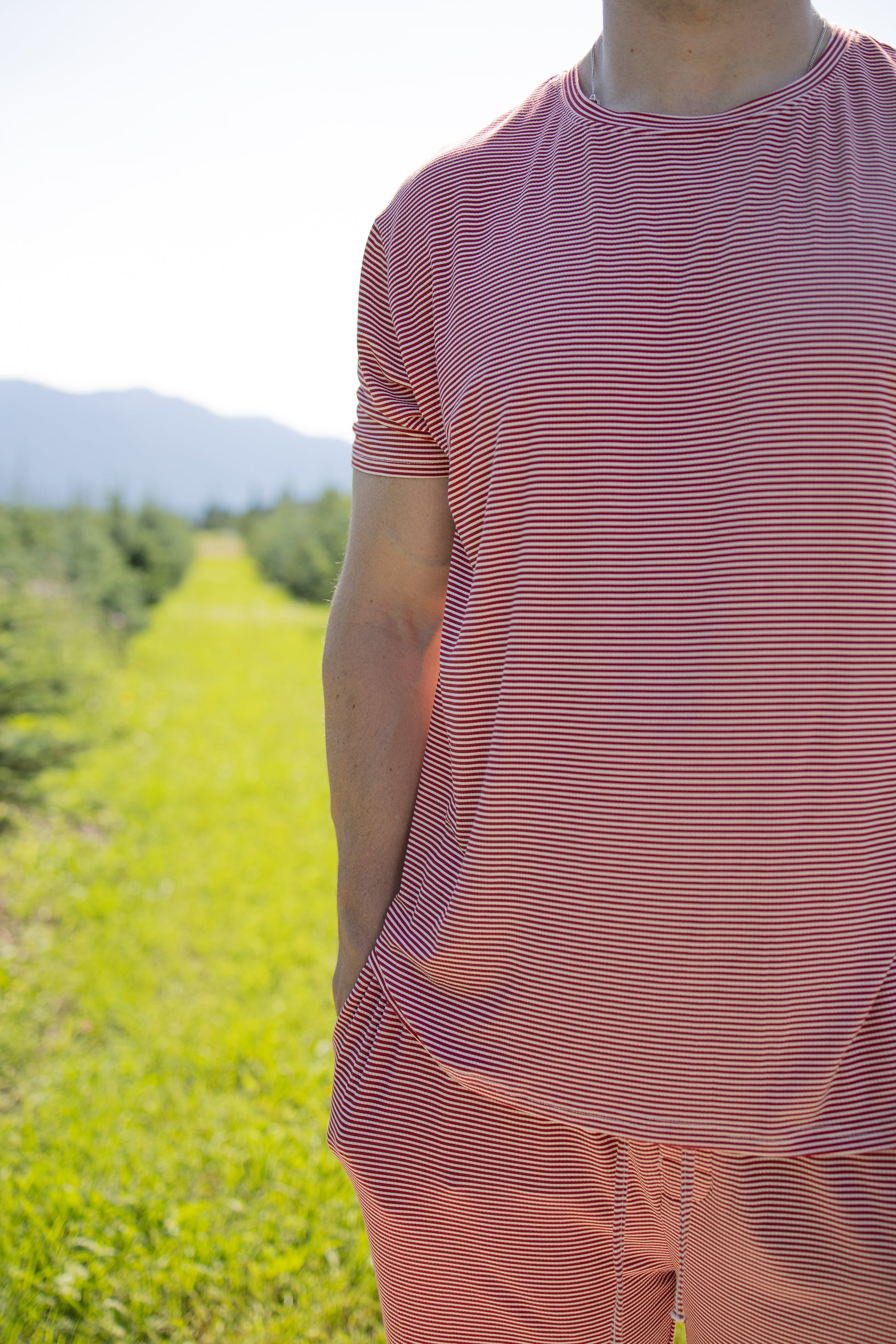 A person wears the forever french baby Men's Short Sleeve Pajamas in Peppermint Stripe Ribbed while standing in a sunlit grassy field with trees and mountains visible in the background.