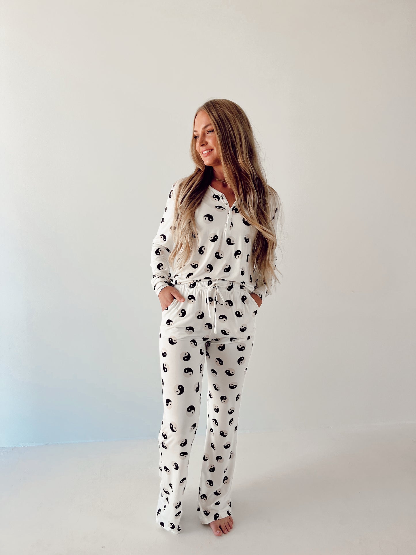 A woman with long hair stands against a plain white background, wearing the Women's Bamboo Pajamas in Black & White Yin Yang from forever French baby. She has her hands in her pockets and is smiling, looking off to the side.