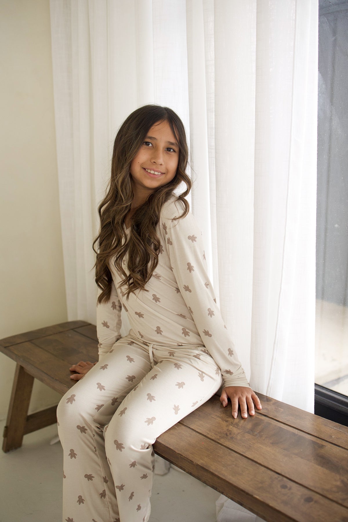 A girl wearing beige long-sleeve Youth Flare Bamboo Pajamas | Gingersnap with a repeating gingerbread man pattern sitting on a wooden bench.