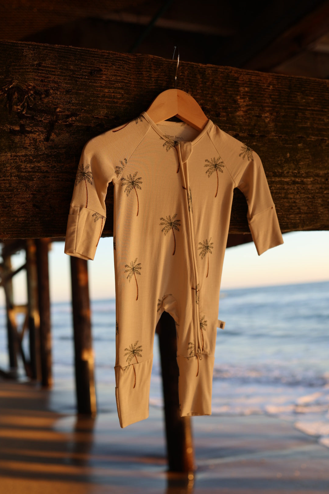 The Bamboo Zip Pajamas | Palm Tree by forever french baby, a hypoallergenic beige romper with palm tree print, hangs on a wooden hanger under a pier at the beach with ocean waves and sunlight in the background.