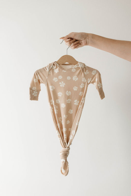 A hand holds the forever french baby Lazy Daisy Bamboo Knotted Gown, featuring a beige color with white daisy patterns and a knotted bottom, set against a plain white background.