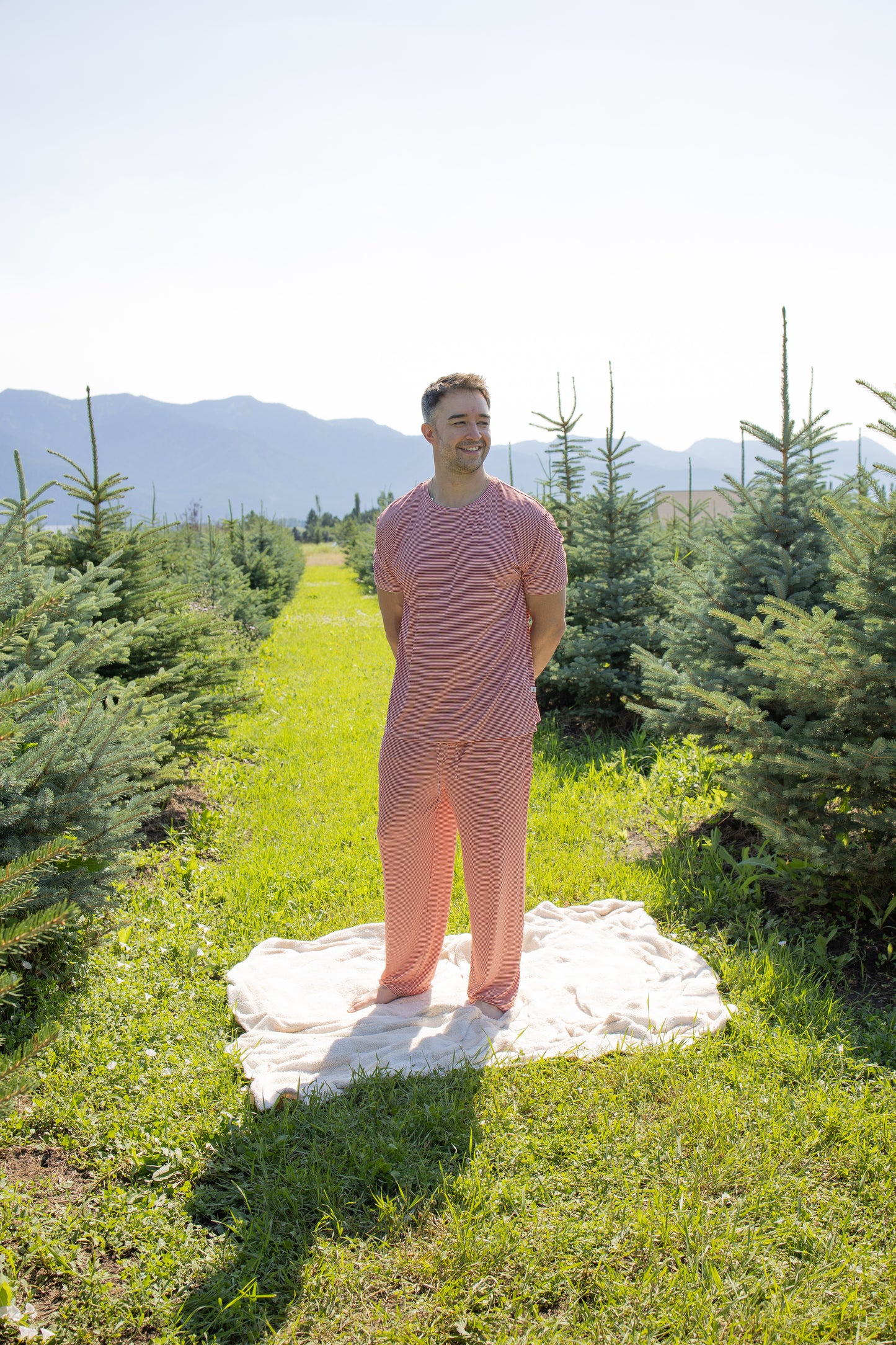 A person wearing the forever french baby Men's Short Sleeve Pajamas in Peppermint Stripe Ribbed stands on a white blanket in a field of small evergreen trees, with mountains and a clear sky in the background.