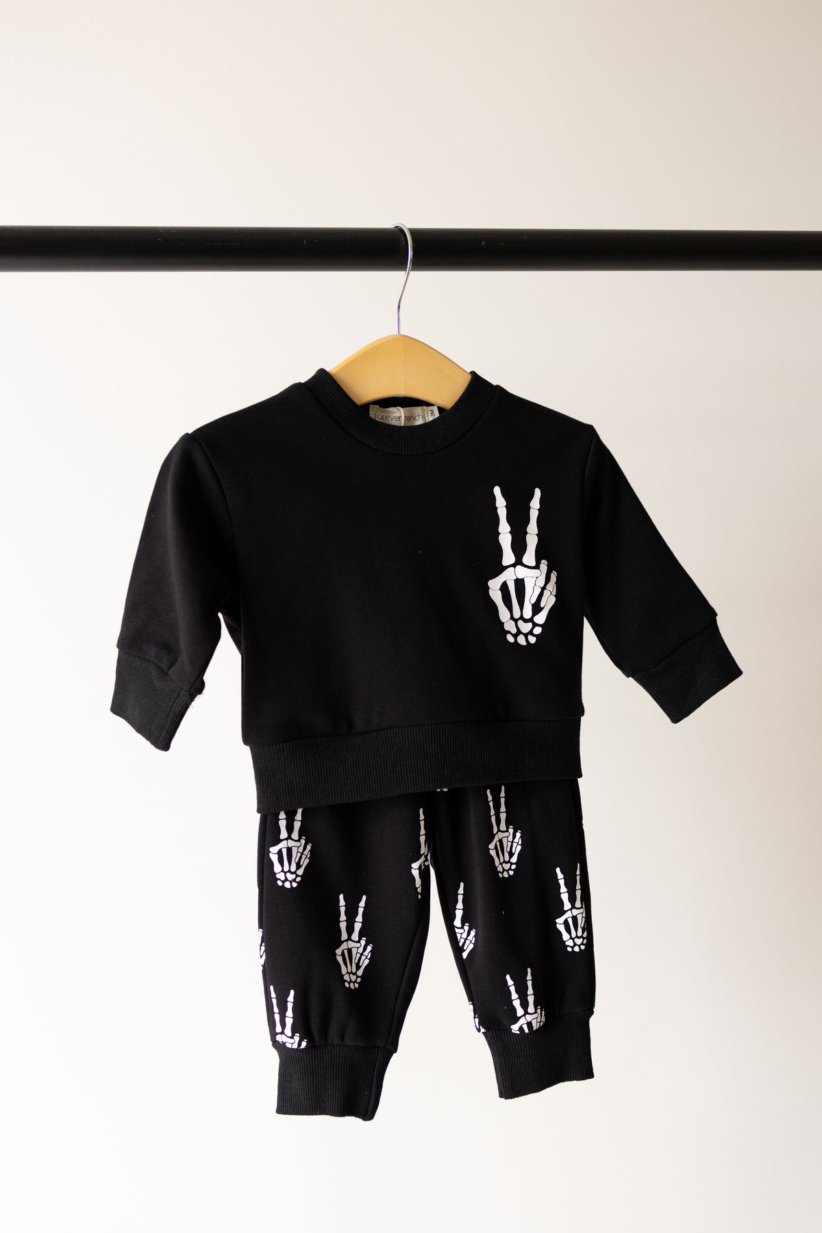 The Child Sweat Set | Midnight Skelly by forever french baby, a black children’s sweat set adorned with white skeleton hand peace signs on both the sweatshirt and pants—perfect for everyday adventures—is displayed on a wooden hanger against a plain light background.