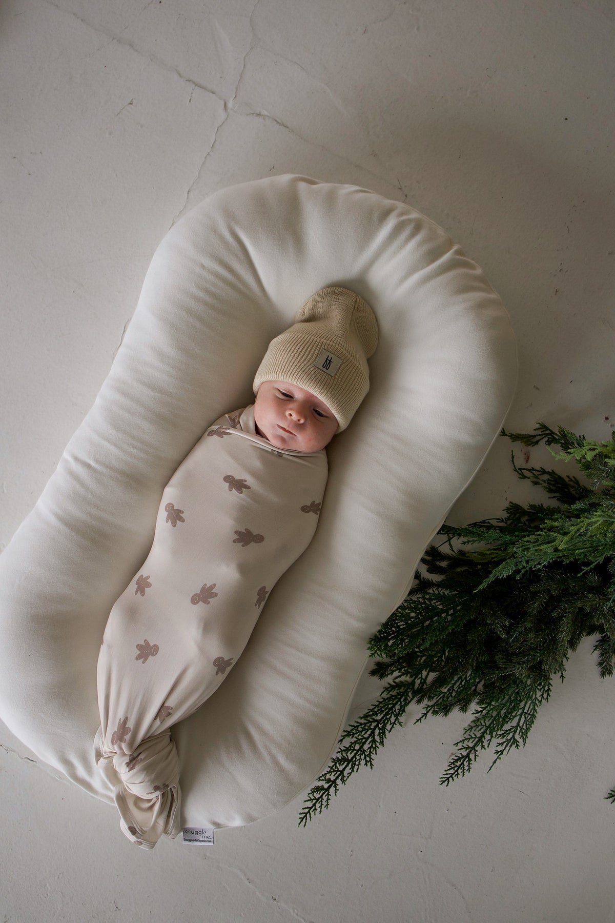A baby wearing a tan beanie lies in a lounger, wrapped in a Bamboo Swaddle | Gingersnap featuring a gingerbread man print.
