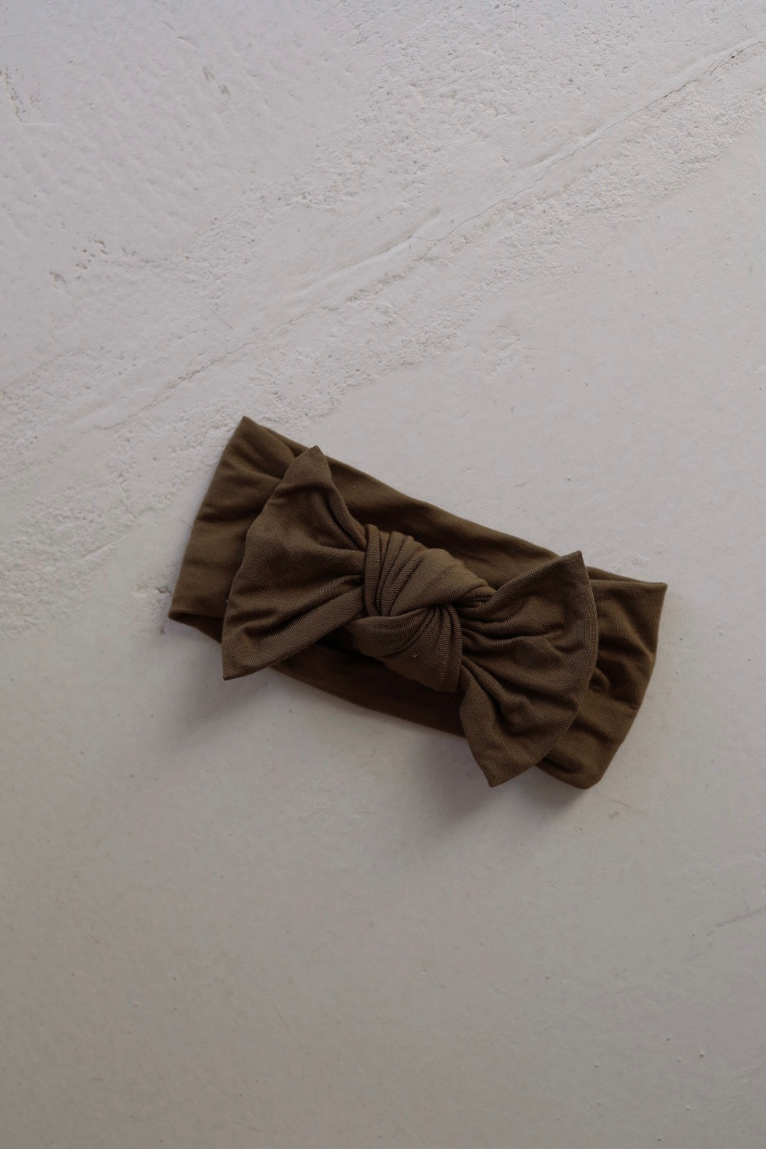 The forever french baby Bamboo Head Wrap in olive green, featuring a large, adjustable bow, is displayed on a light-colored, textured surface.