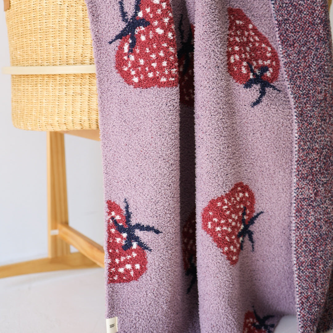The forever french baby Plush Blanket | Matilda, a velvet-soft purple microfiber blanket with large red strawberry patterns, is draped over a woven basket on wooden legs. Oeko-Tex certified for peace of mind.