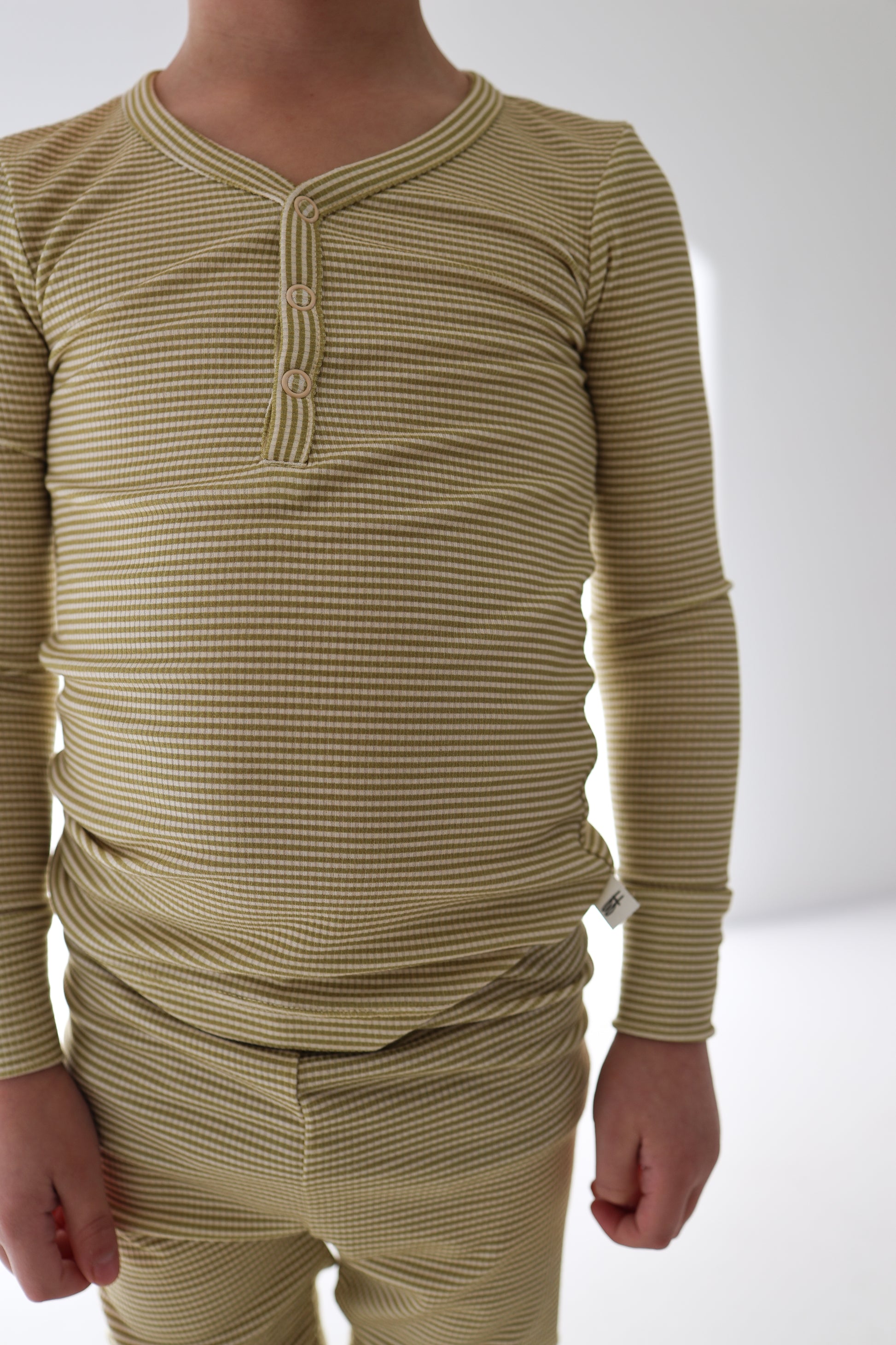 A young child in forever french baby Bamboo Two Piece Pajamas | Willow Stripe Ribbed—a yellow-and-white striped, long-sleeve henley top with matching pants—stands indoors against a plain white background.