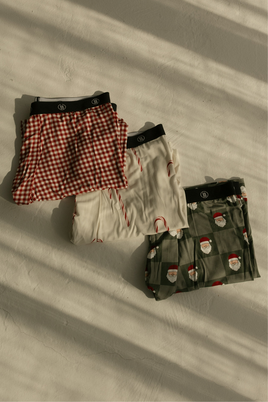 Three pairs of forever french baby Men's Bamboo Boxers in Red Gingham, Candy Cane, and Santa Checkerboard prints lie folded with black waistbands on a light surface, sunlight highlighting their soft, breathable fabric.