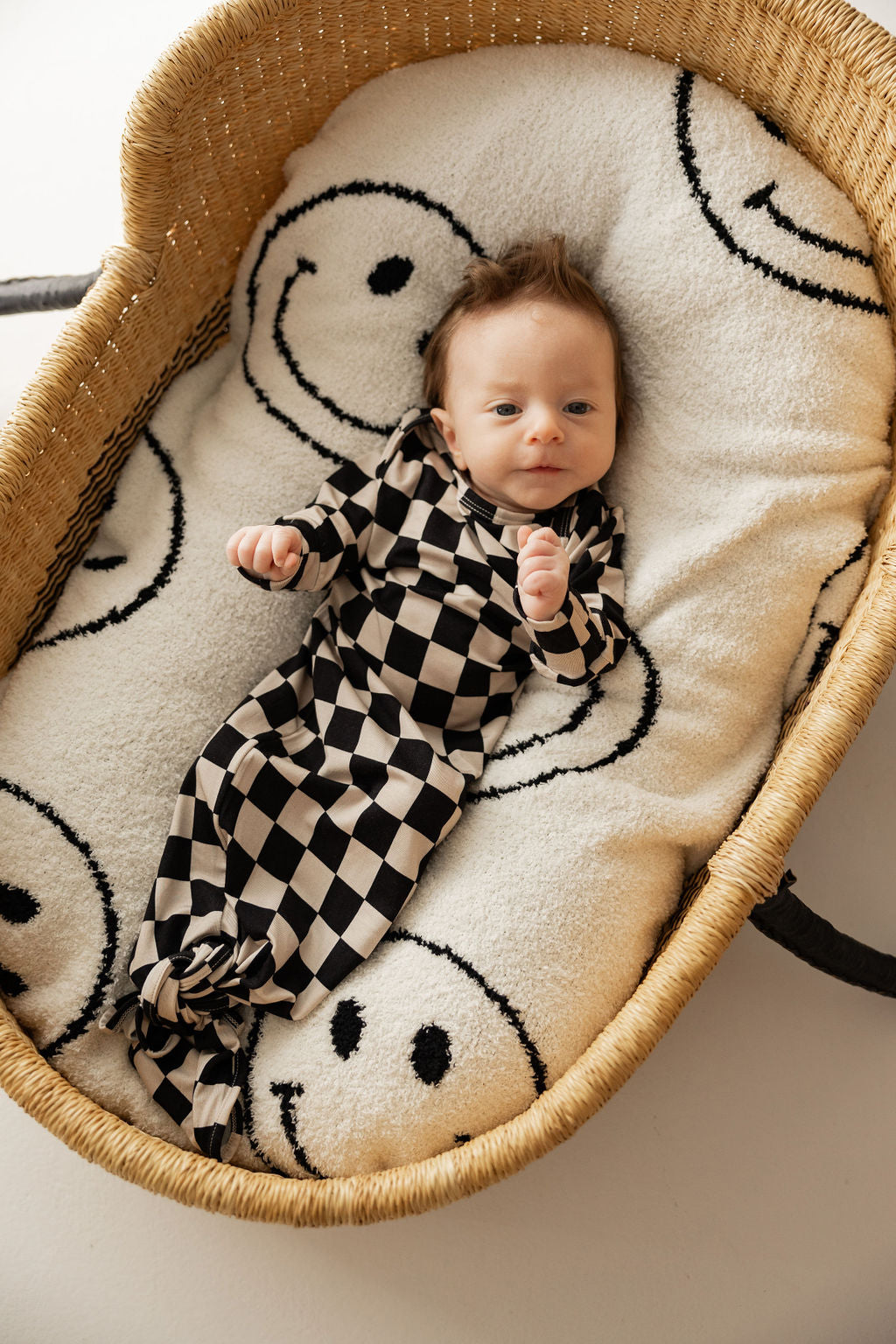 A baby lying in a wicker bassinet lined with a blanket featuring black and white smiley faces is wearing a Knotted Bamboo Gown in Black Checkered from the brand forever french baby, looking relaxed.