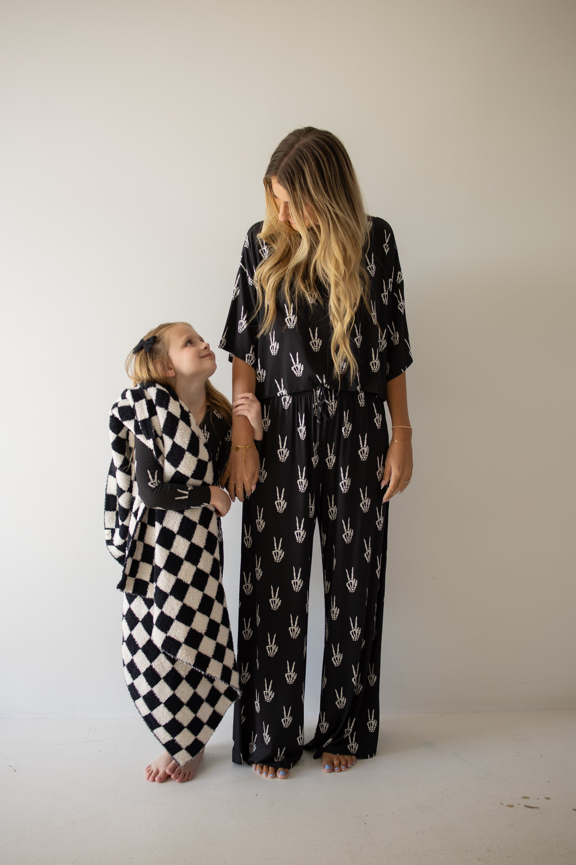 A woman and a young girl stand against a plain wall, smiling at each other. The woman wears the forever french baby Women’s Bamboo Crop Tee Lounge Set in the Midnight Skelly print.