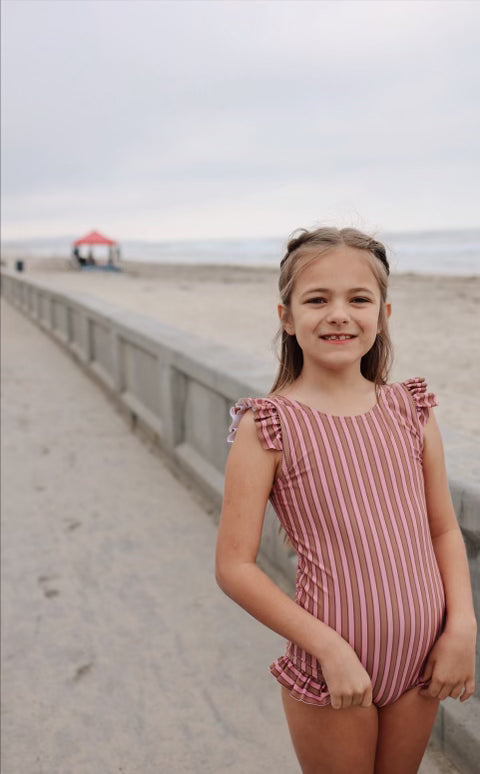 A young girl with braided hair stands on a sandy beach near a concrete railing. She is wearing the "One Piece Girls Swim Suit | Boardwalk" by forever french baby, featuring pink and white stripes with ruffled shoulder straps and offering UPF 50+ protection, while smiling at the camera. The sky is overcast, and there is a tent visible in the background.