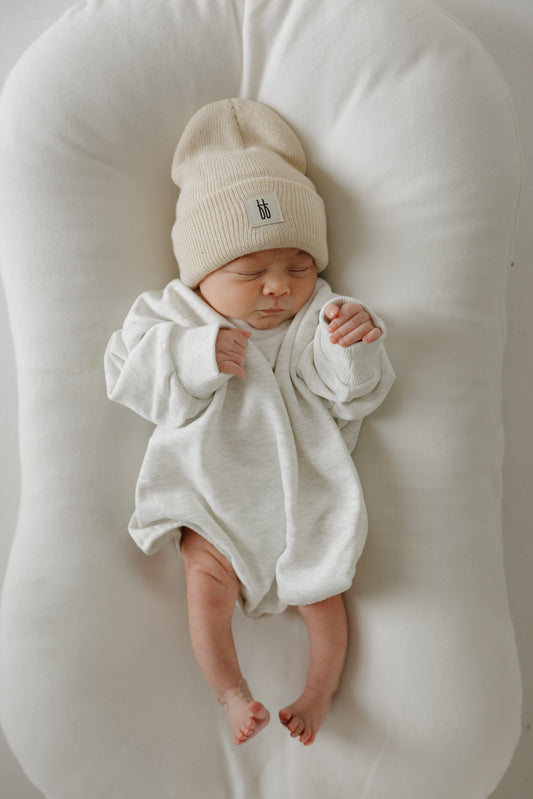 A newborn sleeps peacefully on a soft white cushion, dressed in the forever french baby Sweatshirt Romper | Heather Grey, with a beige knit hat and light gray onesie—perfect for stylish newborn clothing lovers.