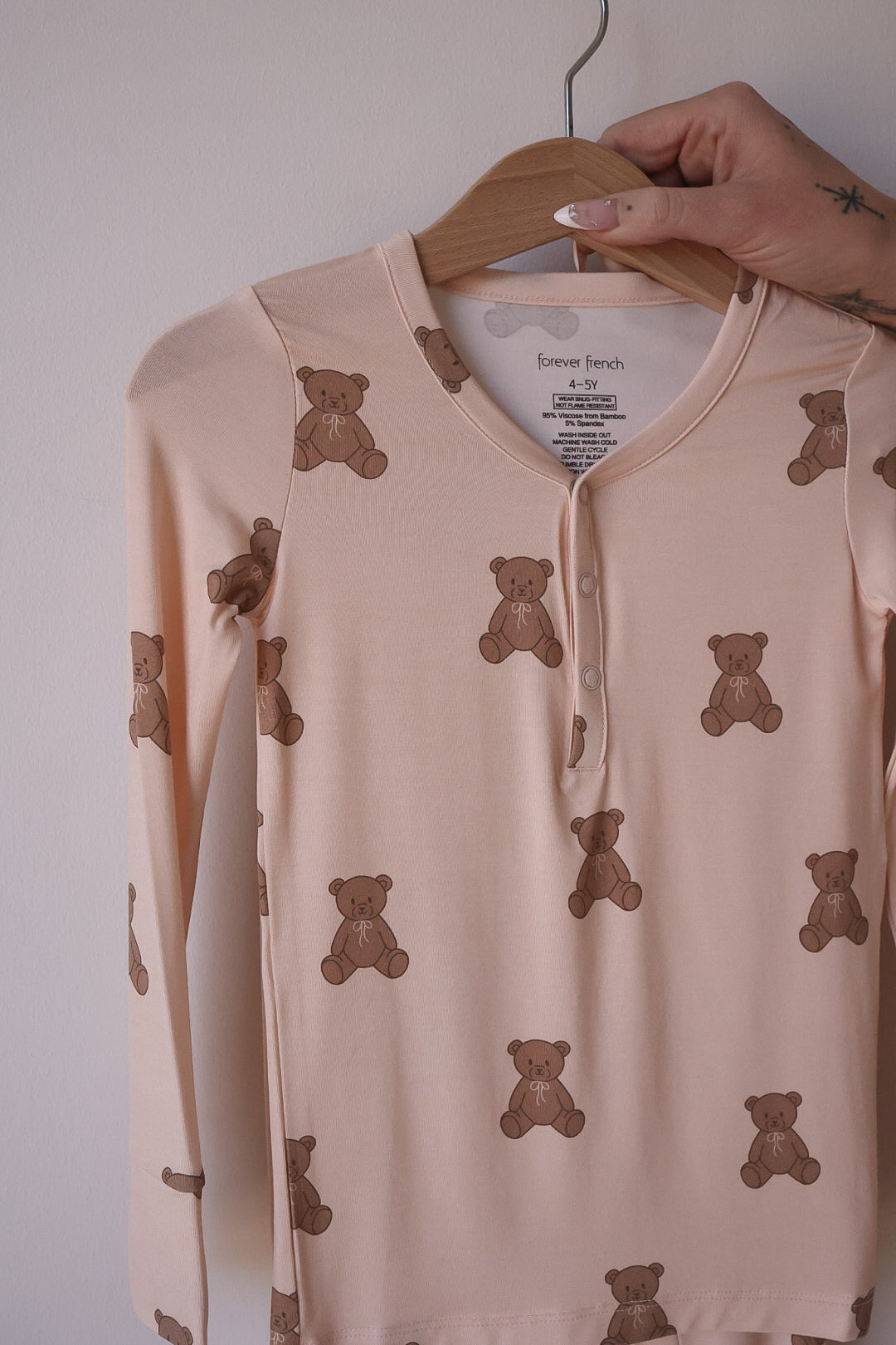 The Bamboo Two Piece Pajamas | Lottie by forever french baby, featuring beige long sleeves and brown teddy bear prints, hang on a wooden hanger held by a tattooed hand against a light-colored wall.