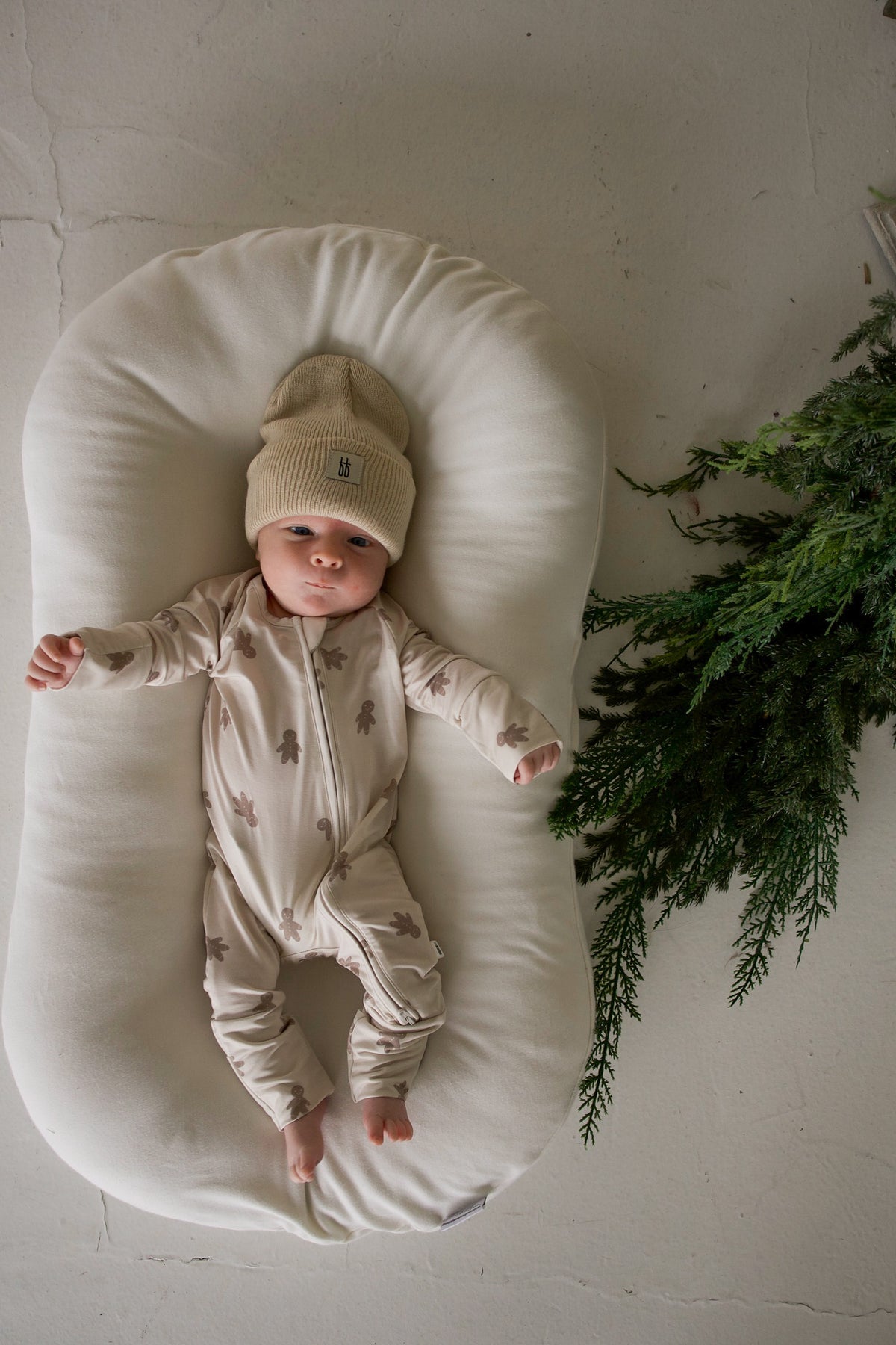 A baby in a beige beanie and Bamboo Zip Pajamas | Gingersnap, lying in a cream-colored baby lounger next to a pine branch.
