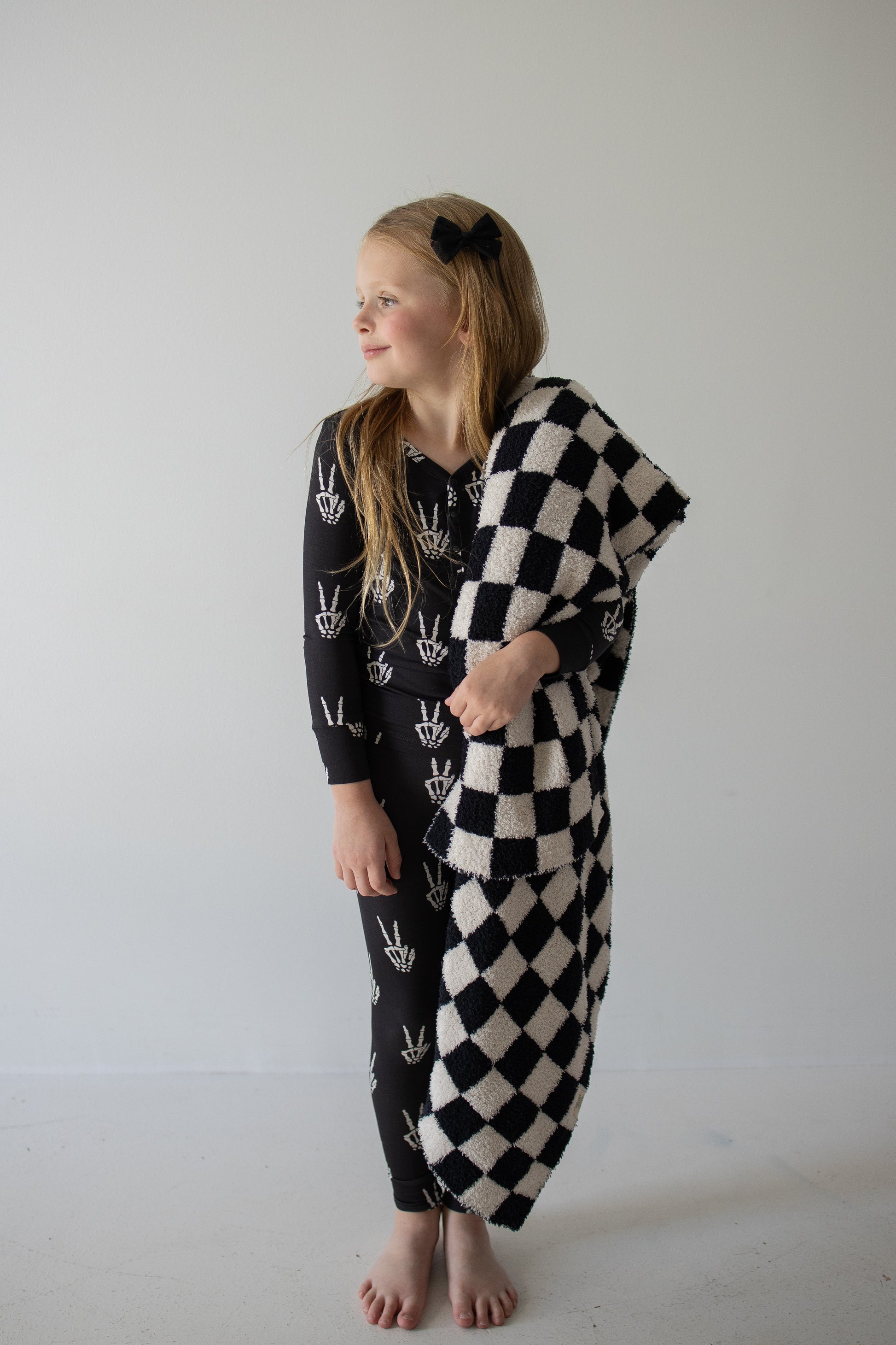 A young girl with long blonde hair stands barefoot against a plain white wall, smiling and looking to her left. She’s dressed in forever french baby’s Bamboo Two Piece Pajamas in the Midnight Skelly print—breathable, stylish sleepwear designed for all-night comfort. She holds a black and white checkered blanket draped over one arm.