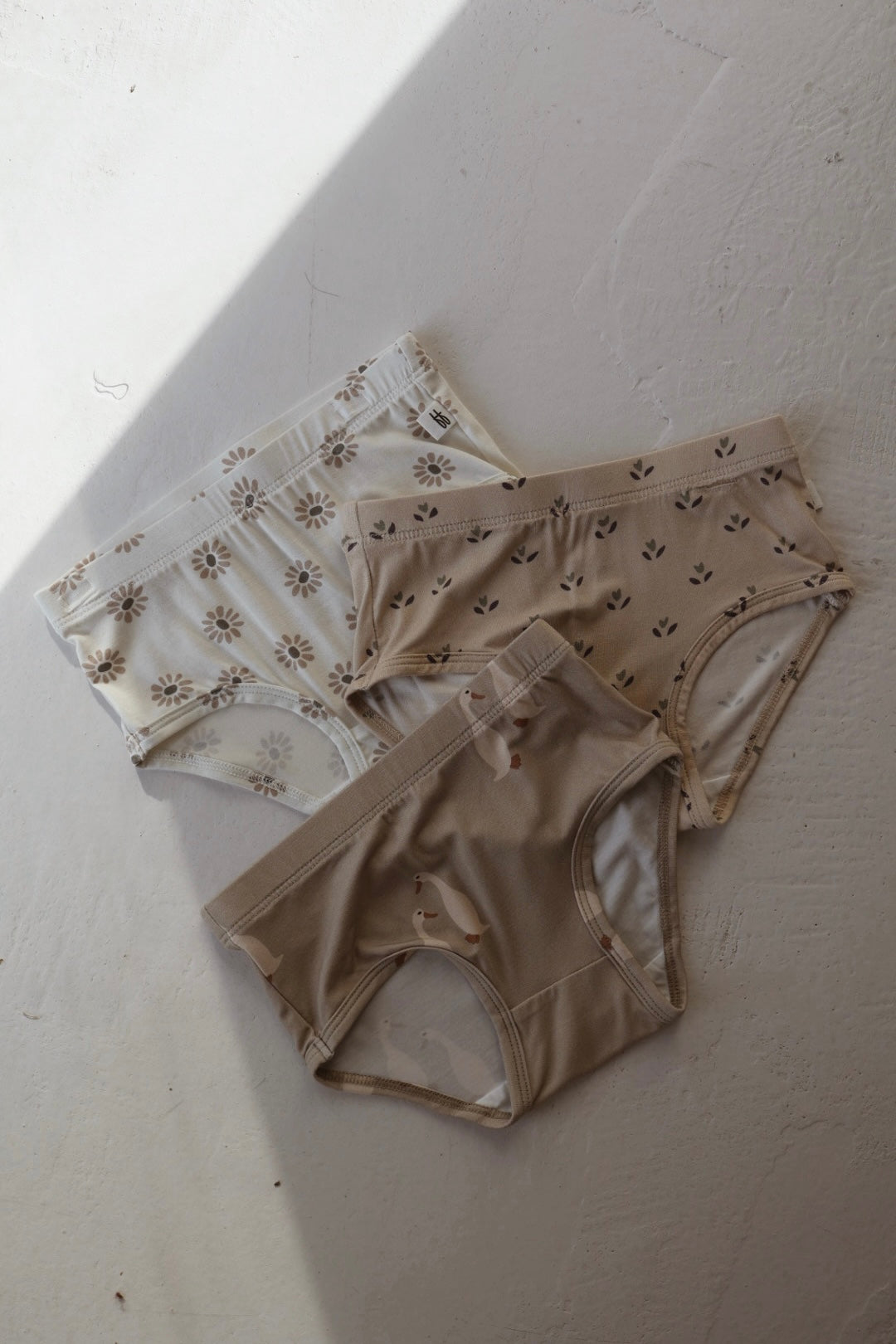 Three pairs of forever french baby Girl’s Bamboo Briefs (3 Pack) in Flora, Silly Goose & Daisy Fields prints are displayed on a light, textured surface with partial shadow. Hypo-allergenic and in neutral tones with fun patterns.