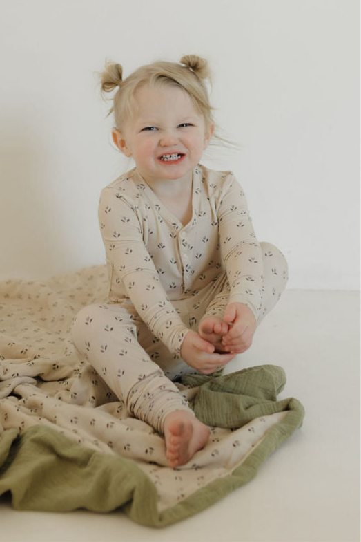 A smiling toddler with light hair in pigtails sits on a cream and green blanket, wearing forever french baby's Bamboo Two Piece Pajamas | Flora, holding one foot and looking at the camera.
