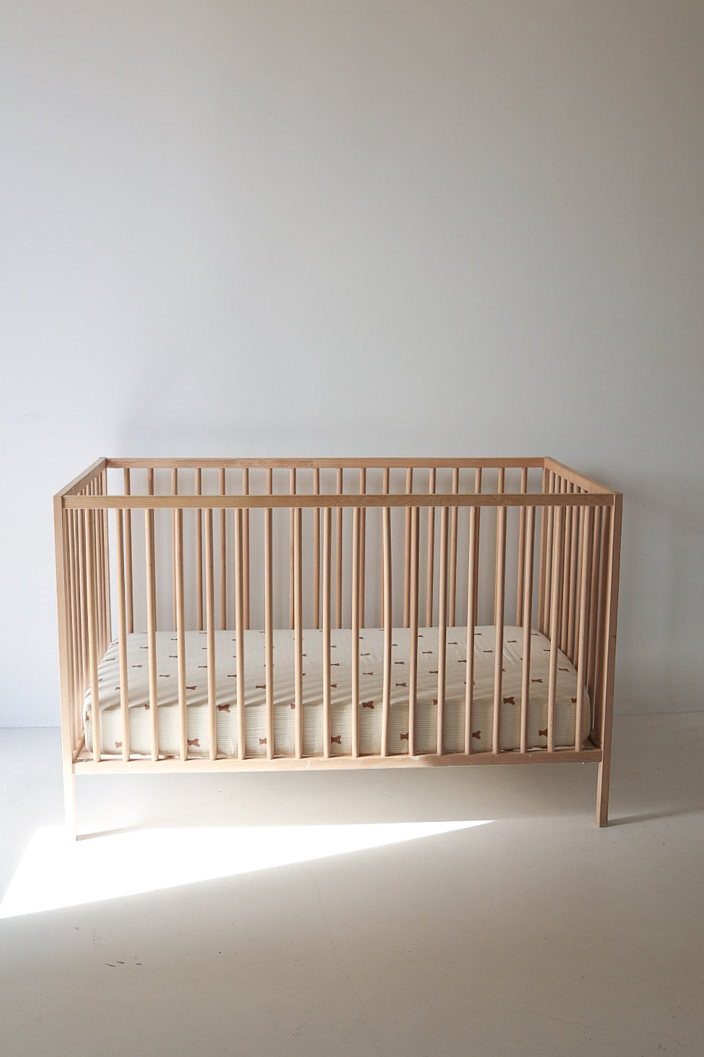 A minimalist wooden crib with vertical slats features the forever french baby Muslin Crib Sheet | Teddy, its subtle light pattern standing out against a plain white wall in a bright, empty room.