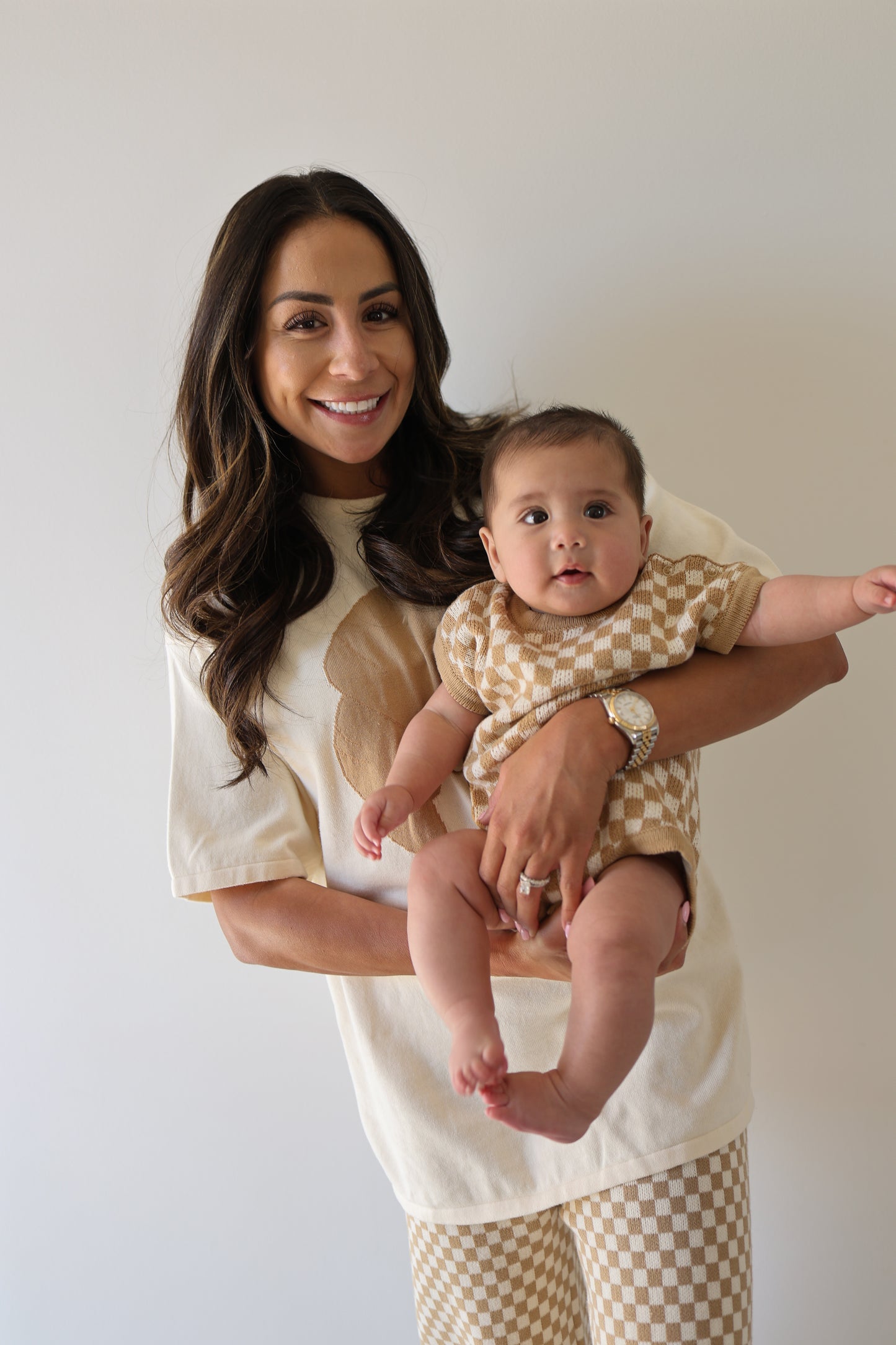 A smiling woman with long brown hair holds a baby wearing the forever french baby Knit Romper in Latte Check. She wears a light shirt and matching checkered pants, standing against a plain white background.