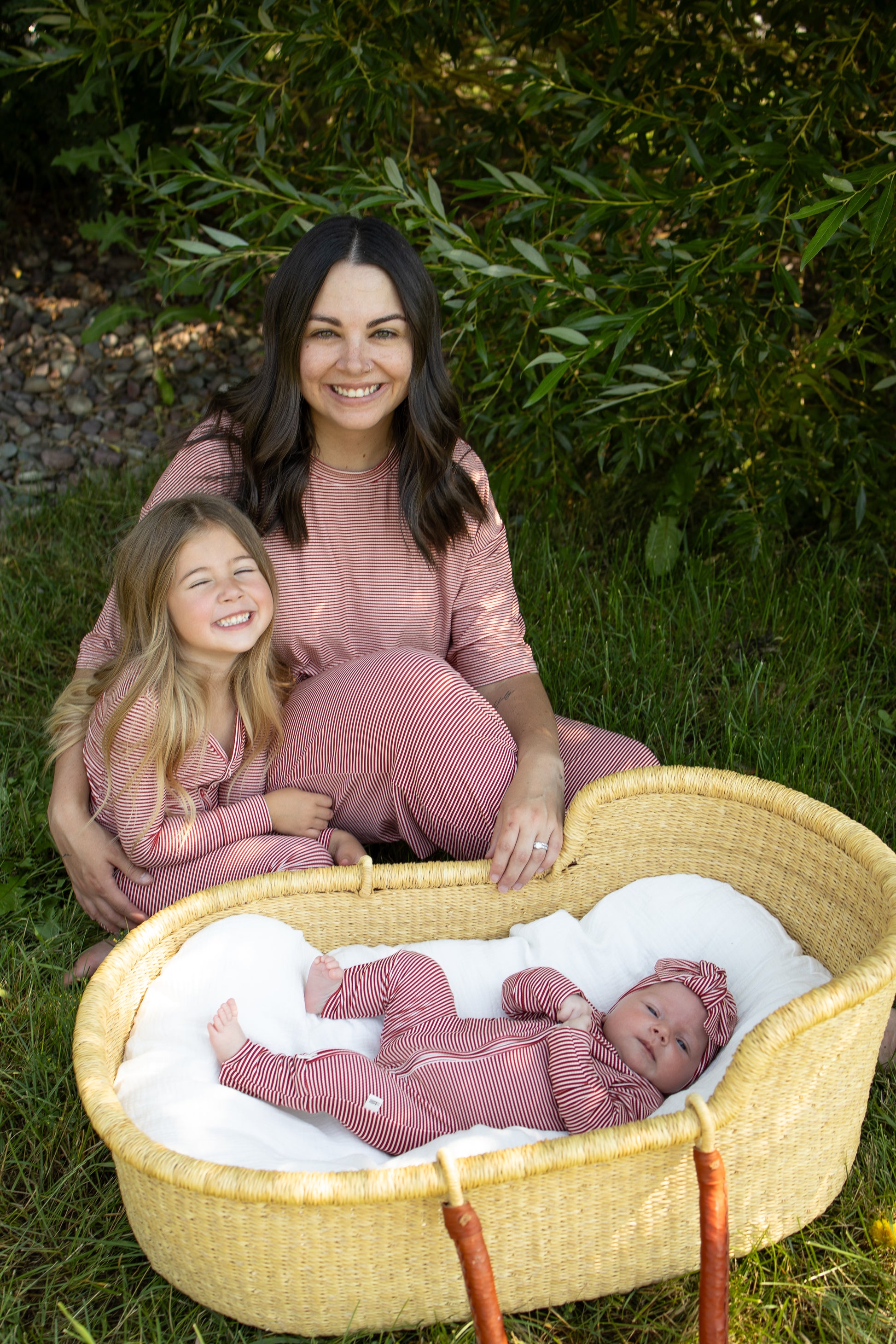 A woman and a young girl sit on the grass, both smiling and wearing matching forever french baby Women's Boxy Tee Lounge Set in Peppermint Stripe Ribbed next to a baby in a wicker basket, also dressed in pink stripes. Their hypo-allergenic sleepwear is perfect for the outdoors, with greenery in the background enhancing this sweet family moment.
