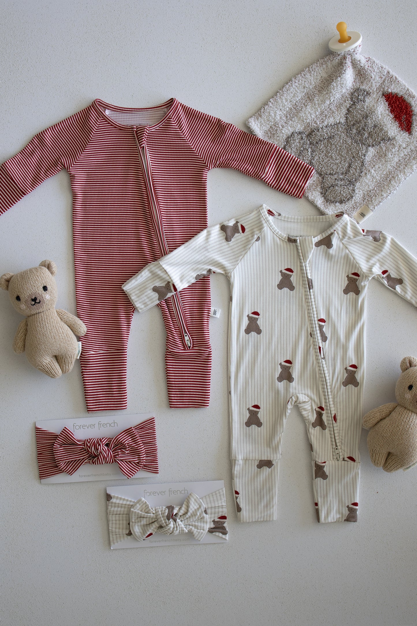 Two baby onesies from forever french baby's Santa Teddy collection—one red striped and one cream with bear print—are displayed alongside the Lovey | Santa Teddy, two teddy bears, a gray washcloth, a pacifier, and two baby headbands on a white surface.