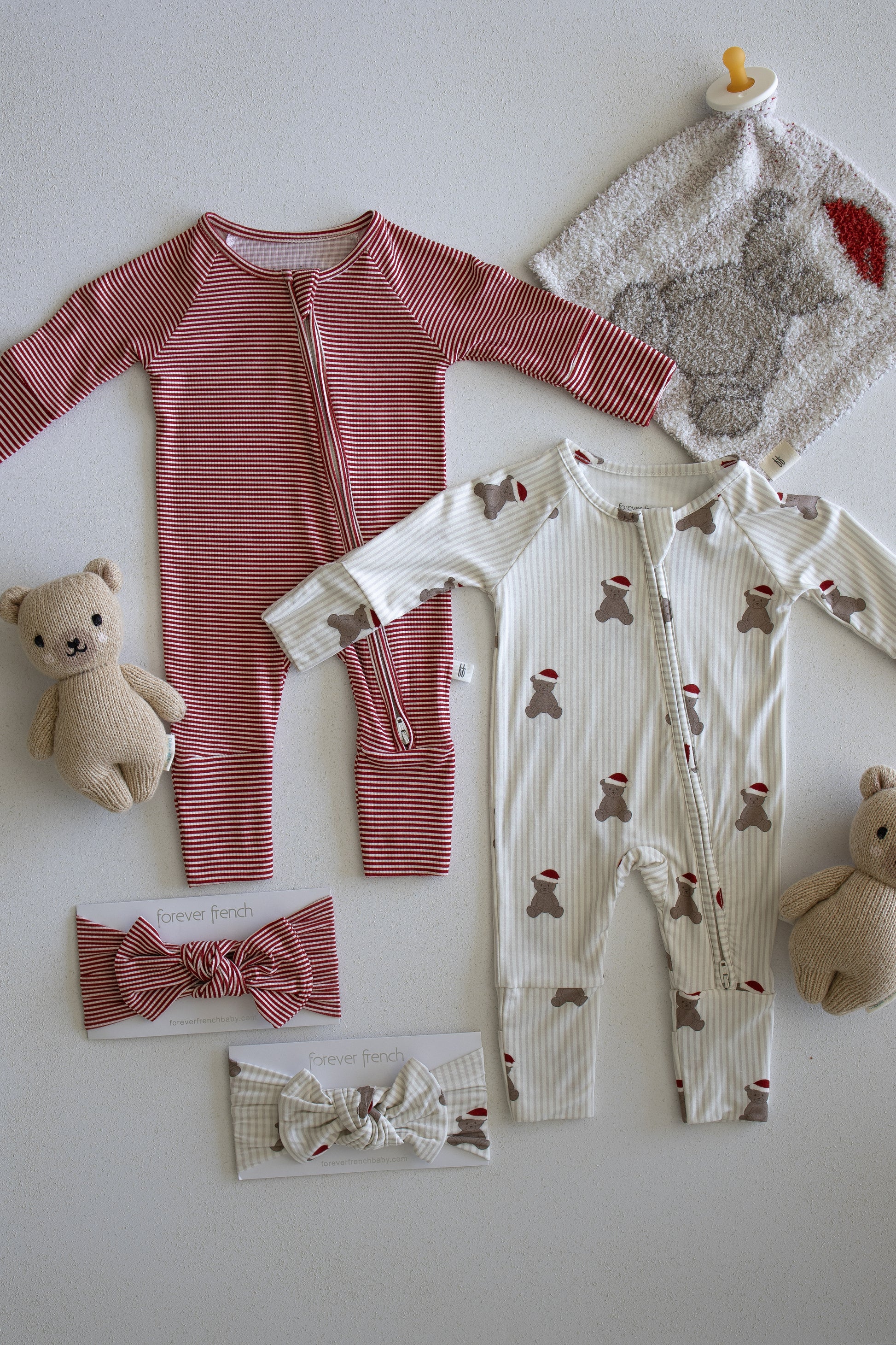 Two baby onesies from forever french baby's Santa Teddy collection—one red striped and one cream with bear print—are displayed alongside the Lovey | Santa Teddy, two teddy bears, a gray washcloth, a pacifier, and two baby headbands on a white surface.