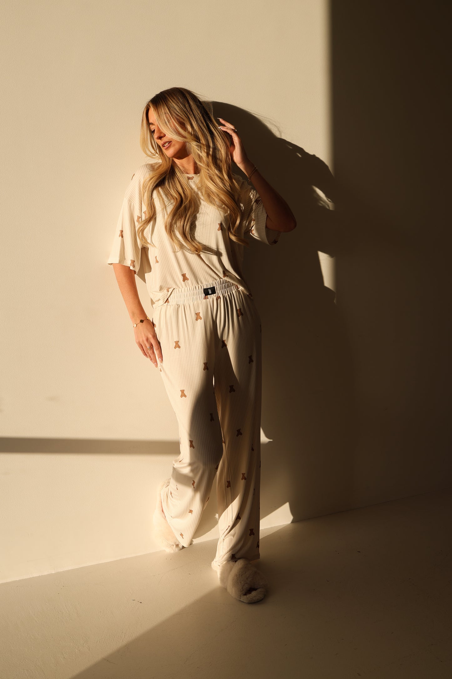 A woman with long blonde hair leans against a sunlit wall, wearing the forever french baby Women's Boxy Tee Lounge Set | Teddy with small patterns and fluffy slippers, as soft shadows are cast behind her.